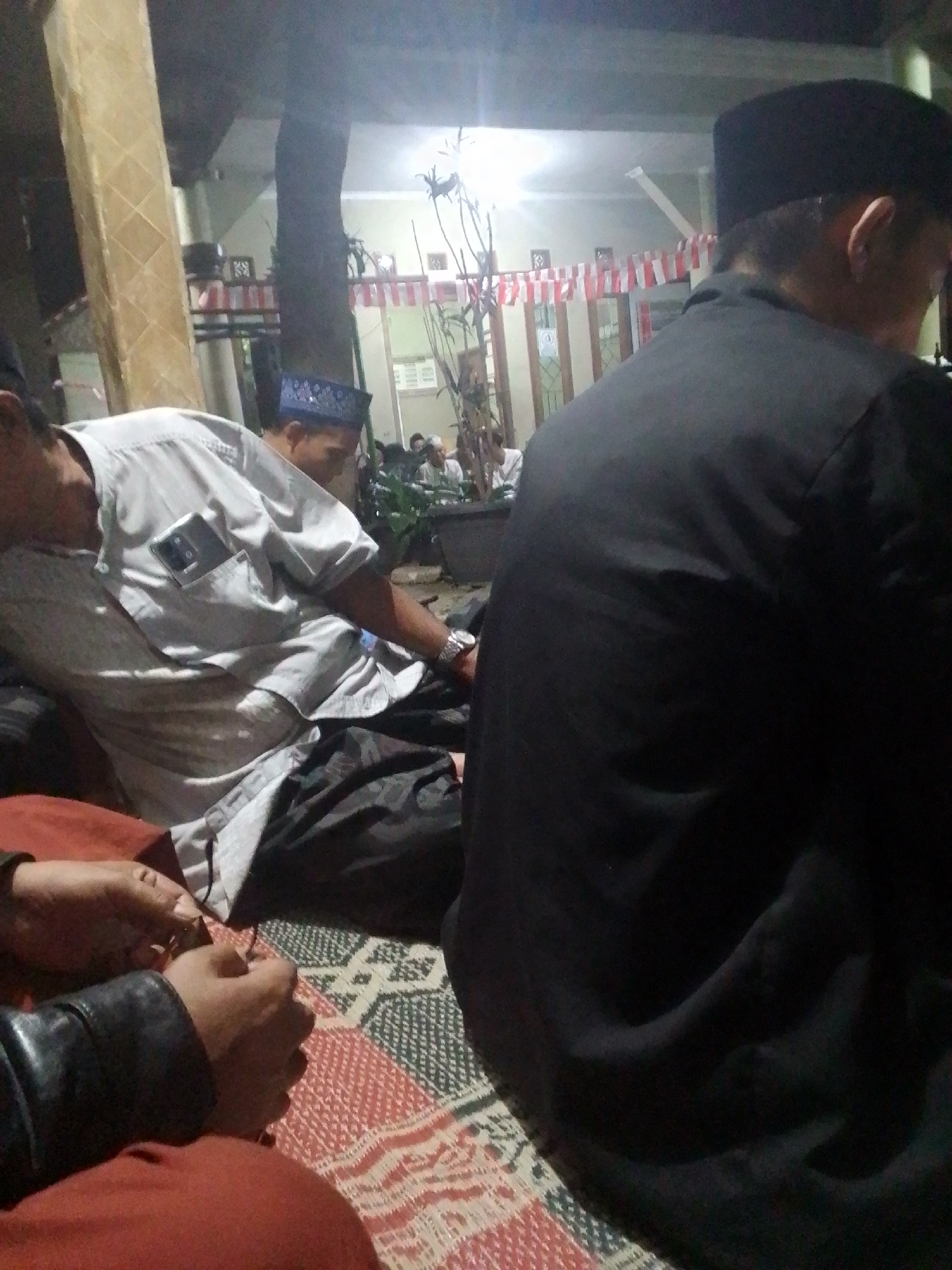 In Sukamanah, Indonesia, an outdoor gathering is depicted at night. Three individuals are visible. In the foreground, a person wearing a black skullcap and black shirt is seated, with their back to the camera. To the left, a man in a light-colored shirt with a phone in his chest pocket and a black sarong is leaning back, appearing to be asleep. Behind him, another man in a blue peci hat is also seated. Red and white Indonesian flags are strung across the background, suggesting a celebration or event. The ground is covered with a patterned mat.