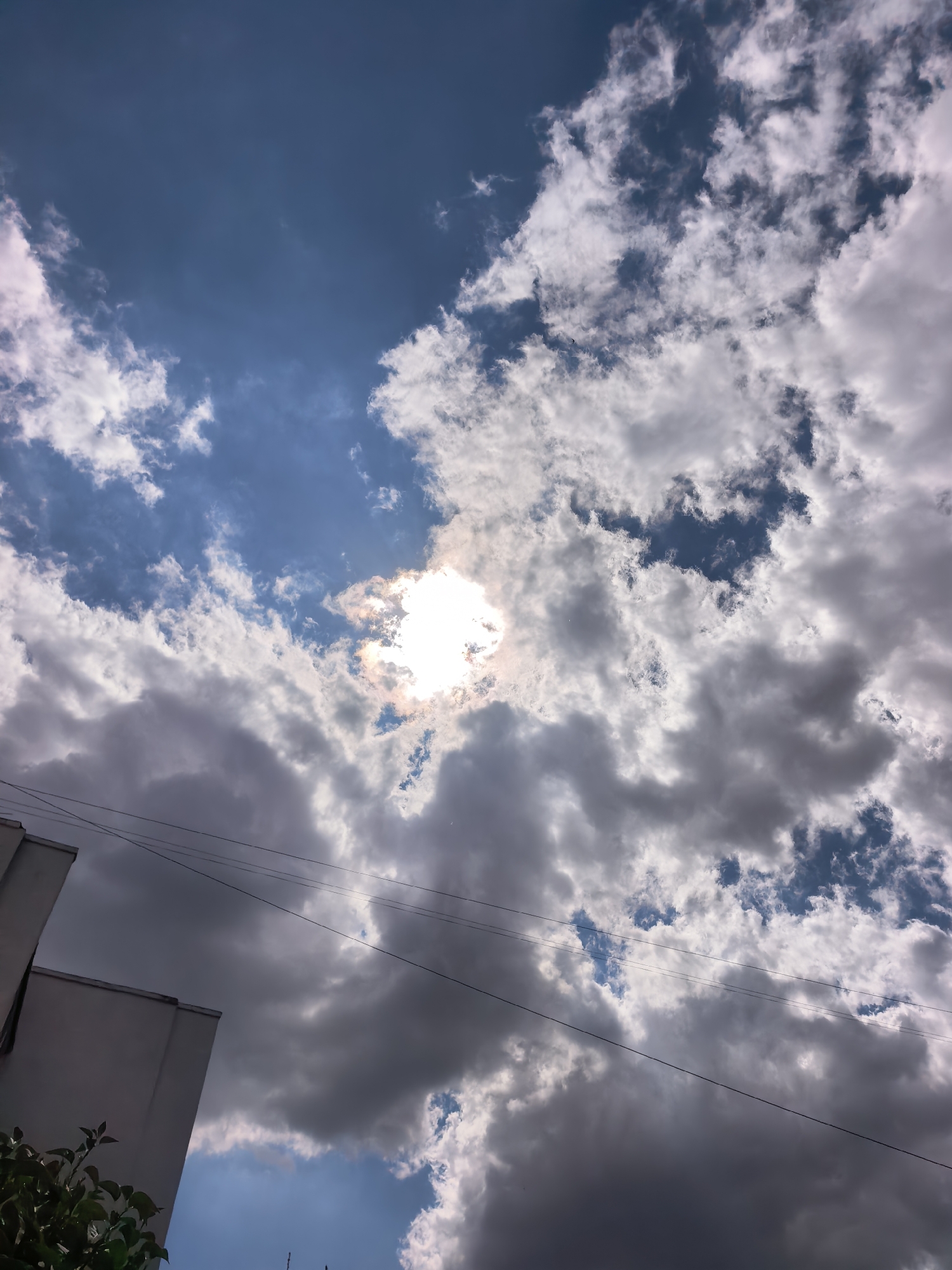 The image captures a low-angle view of a cloudy sky above a city. The sun is partially obscured by thick, grey clouds, with patches of bright blue sky visible in between. The clouds vary in texture and density, with some appearing wispy and light, while others are dark and heavy. Power lines are strung across the sky, adding a man-made element to the natural scene. In the lower left corner, a portion of a building, possibly a wall of a light-colored structure, is visible, along with some dark green foliage. The overall impression is of a dramatic sky with a hint of urban landscape. The location is identified as Sterlitamak, Russia.