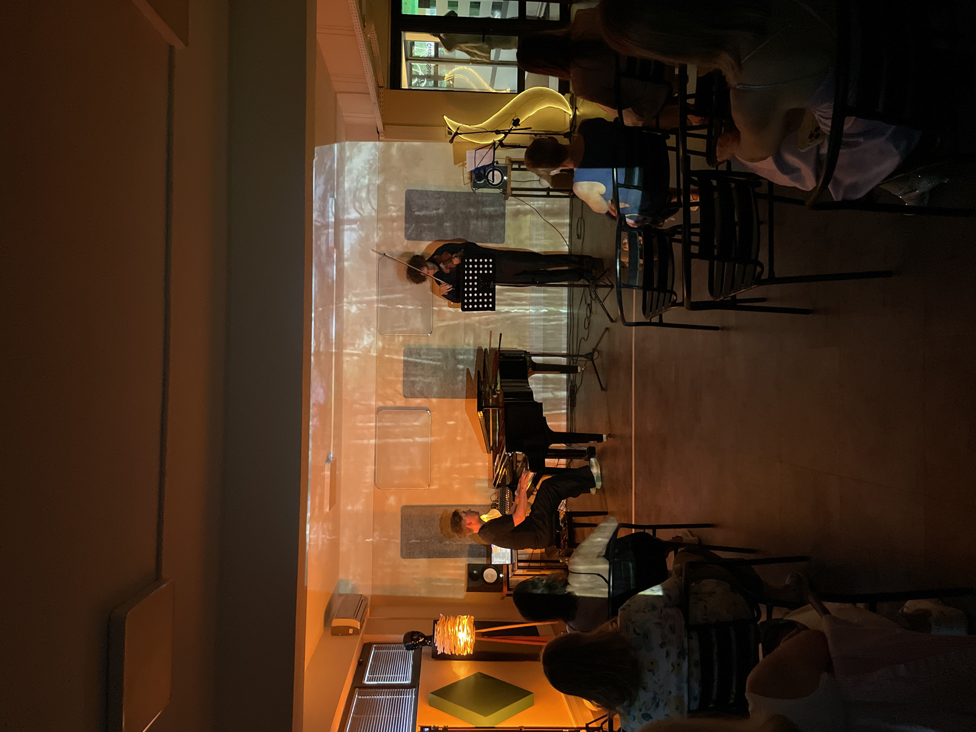 The image shows a dimly lit room in Tivat, Montenegro, likely a bar or small performance venue, set up for a musical performance. A violinist and a pianist are positioned in the center of the room, playing music. Both have music stands in front of them. The pianist sits at a black upright piano. An audience sits in chairs, facing the musicians. The walls are illuminated with what appears to be a projected image. A modern decorative lamp is visible in the foreground.