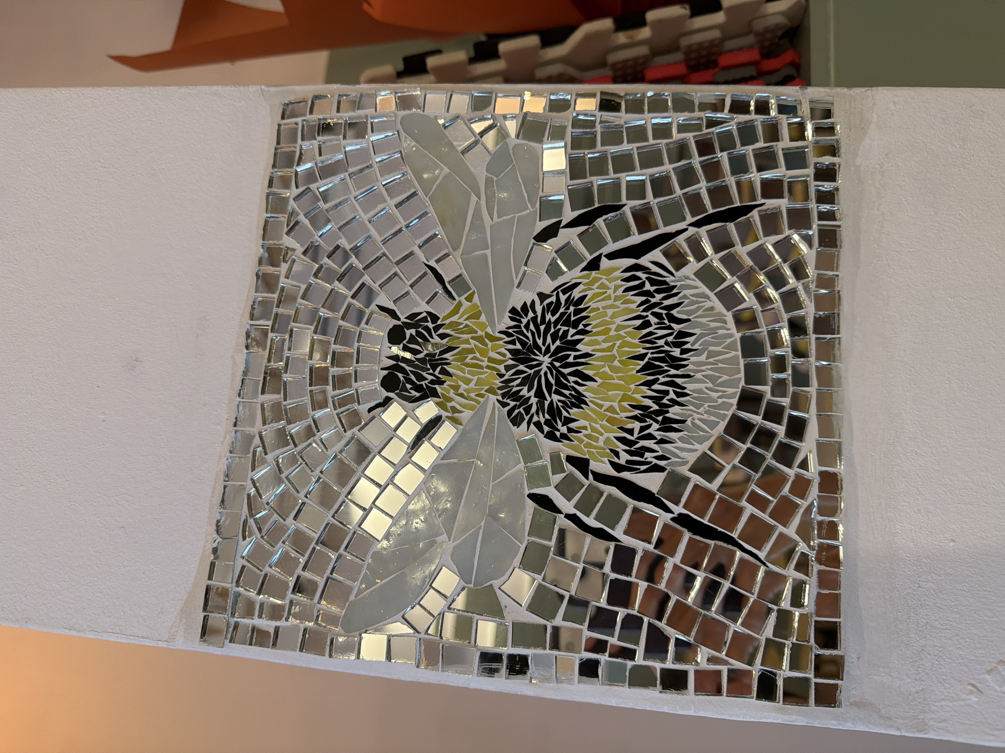 The image shows a mosaic depicting a bee. The bee is made up of small, square tiles in shades of yellow, black, and white. The wings are white, and the body has a striped pattern. The background is composed of reflective, square tiles. The mosaic is mounted on a white wall. The presence of the mosaic hints at an artistic or decorative intention. I have no information about the mosaic being located in Budva, Montenegro.