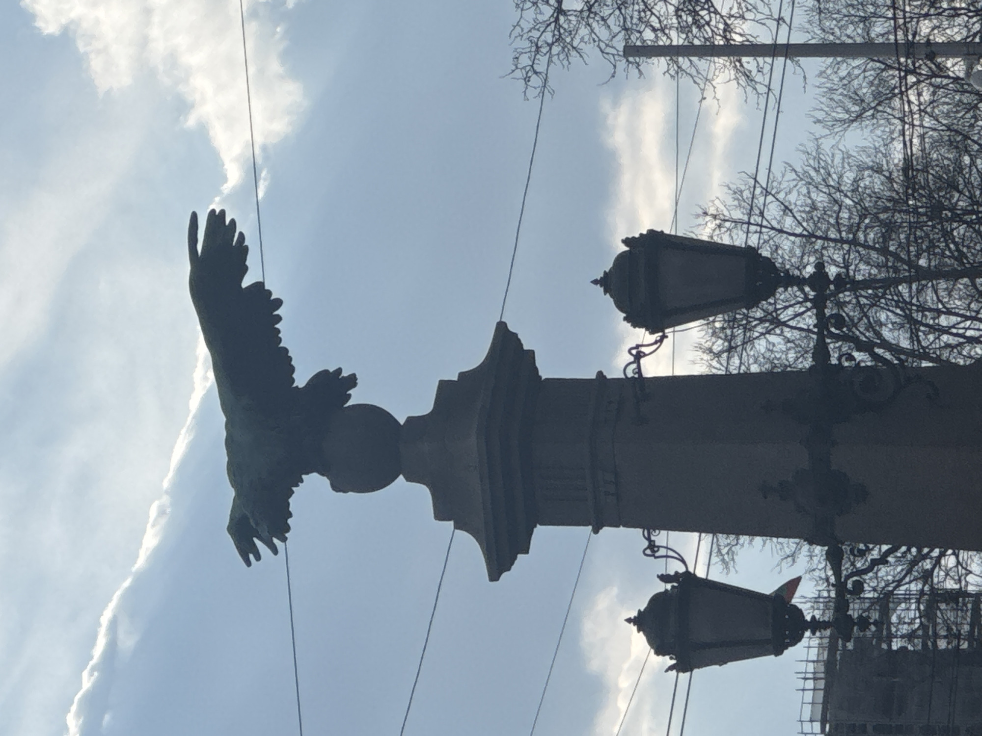 The image displays a low-angle, backlit shot of one of the four bronze eagle statues at Orlov Most (Eagle's Bridge) in Sofia, Bulgaria. The dark, verdigris-patinated eagle is centrally positioned, with its wings fully spread, perched on a spherical element crowning a multi-tiered, fluted stone pillar. Below the eagle, two ornate, dark street lamps with decorative scrollwork are affixed to the pillar, one on each side, their lantern heads detailed with finials. Bare deciduous tree branches are visible, silhouetted against the sky on the right side of the frame. Multiple thin, dark wires (likely overhead power or tram lines) crisscross the sky horizontally. In the distant background, to the left of the central pillar, a structure under construction with visible scaffolding is present. A small, faint national flag with horizontal stripes (consistent with the Bulgarian flag: white, green, red) is discernible near this background structure. The sky is bright blue with scattered white and grey clouds, creating a strong silhouette effect for the foreground objects. No people, specific actions, or interactions are visible.