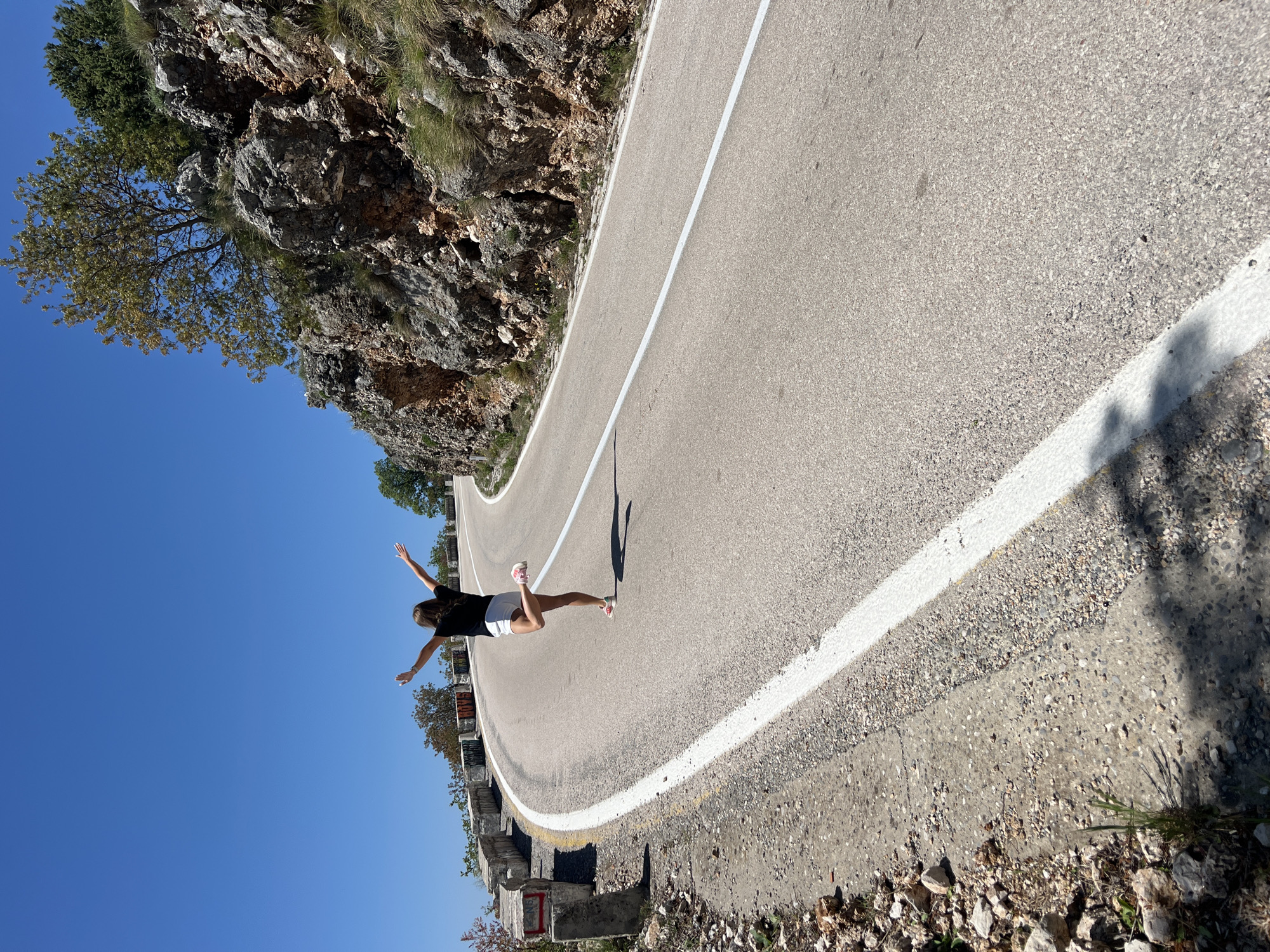 The image, captured in Novoselje, Montenegro, depicts a vibrant outdoor scene on a winding mountain road under a clear blue sky. The road, paved with asphalt, features a distinct solid white line marking its outer edge. It appears to curve gently uphill and to the right from the viewer's perspective.
On the left side of the road, a rugged, rocky cliff face rises, dotted with sparse vegetation, including grass, small bushes, and a few scattered trees. To the right of the road, a lower concrete barrier system separates the asphalt from what appears to be a steep drop-off or open valley, extending towards the clear horizon. Faint, indecipherable markings can be seen on some of the concrete barriers.
A young person, likely female, with long dark hair, is positioned on the road. She is wearing a black t-shirt and white shorts. With bare feet, she strikes a dynamic pose: one leg is bent at the knee and raised slightly, while both arms are outstretched to the sides with palms open. This posture conveys a sense of balance, freedom, or joy, as if in mid-flight or embracing the expansive view. Her elongated shadow stretches across the road in front of her, indicating bright sunlight, likely mid-morning or late afternoon on a clear, warm day.