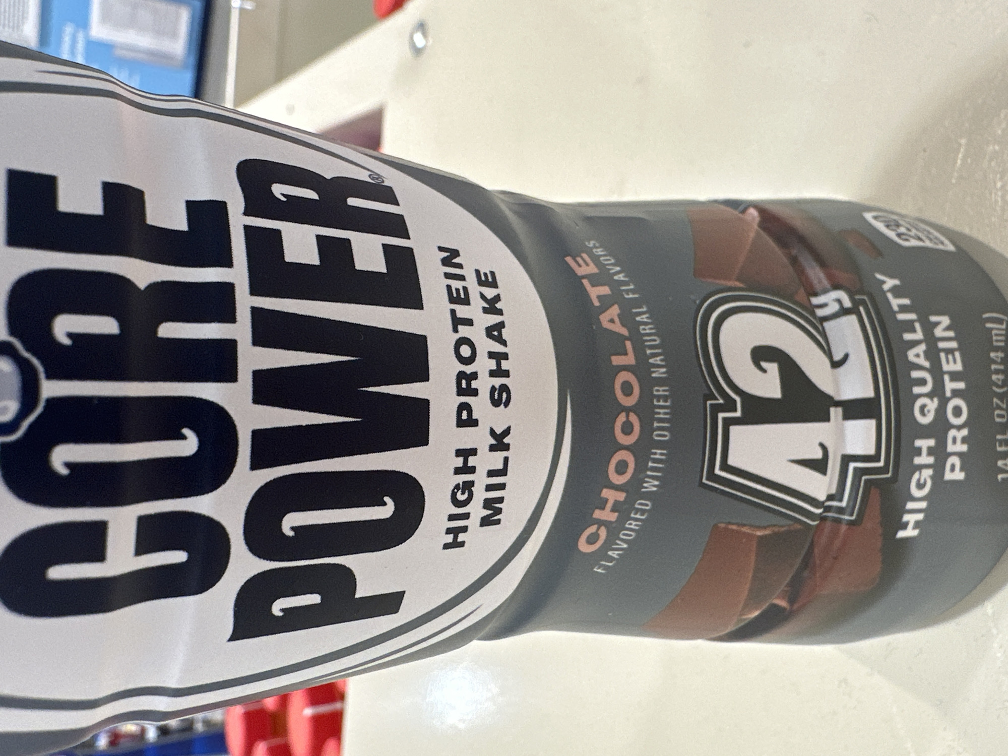 A close-up, slightly angled shot shows the front of a bottle of Core Power High Protein Milk Shake, Chocolate flavor, displayed on a light-colored, speckled surface, likely a countertop. The bottle, primarily gray and white with black text, is the central focus. Large, bold black letters prominently spell out "CORE POWER." Below this, smaller black text reads "HIGH PROTEIN MILK SHAKE." A curved band of darker gray encircles the lower portion of the bottle, featuring the word "CHOCOLATE" in a light pinkish-brown font. Beneath "CHOCOLATE," in smaller white text, is "FLAVORED WITH OTHER NATURAL FLAVORS." Centered on the gray band is a graphic of the number "42" in a stylized font, outlined in black and white. Below the "42" graphic, white text states "HIGH QUALITY PROTEIN." A small white box with the number "230" is visible to the right of the "42" graphic. The background is blurred, showing hints of red objects on the left and a light blue object with a white rectangular barcode on the right. The scene is indoors, with artificial lighting. The image was captured in Camp Hill, United States of America.