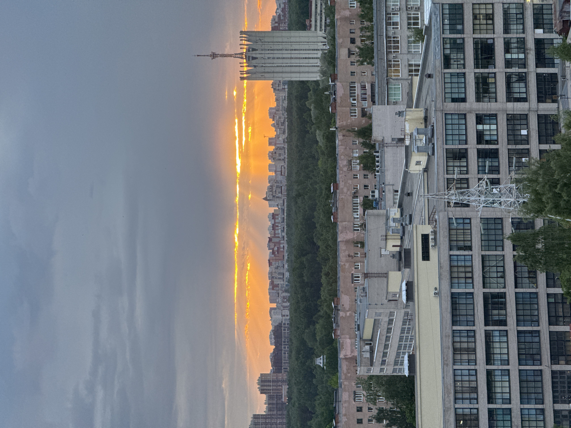 The image shows a city landscape at sunset. The sky is divided, with a dark, cloudy portion on the left and a bright, orange, sunlit portion on the right. In the distance, there are various buildings, including a distinctive tower with a tall antenna, partially silhouetted against the sun. A line of trees stretches vertically down the middle of the image, separating the sunlit and darker parts of the city.
On the right side of the image, there are several apartment buildings with numerous windows. An electrical tower is visible closer to the foreground. The location appears to be Unknown, Unknown.