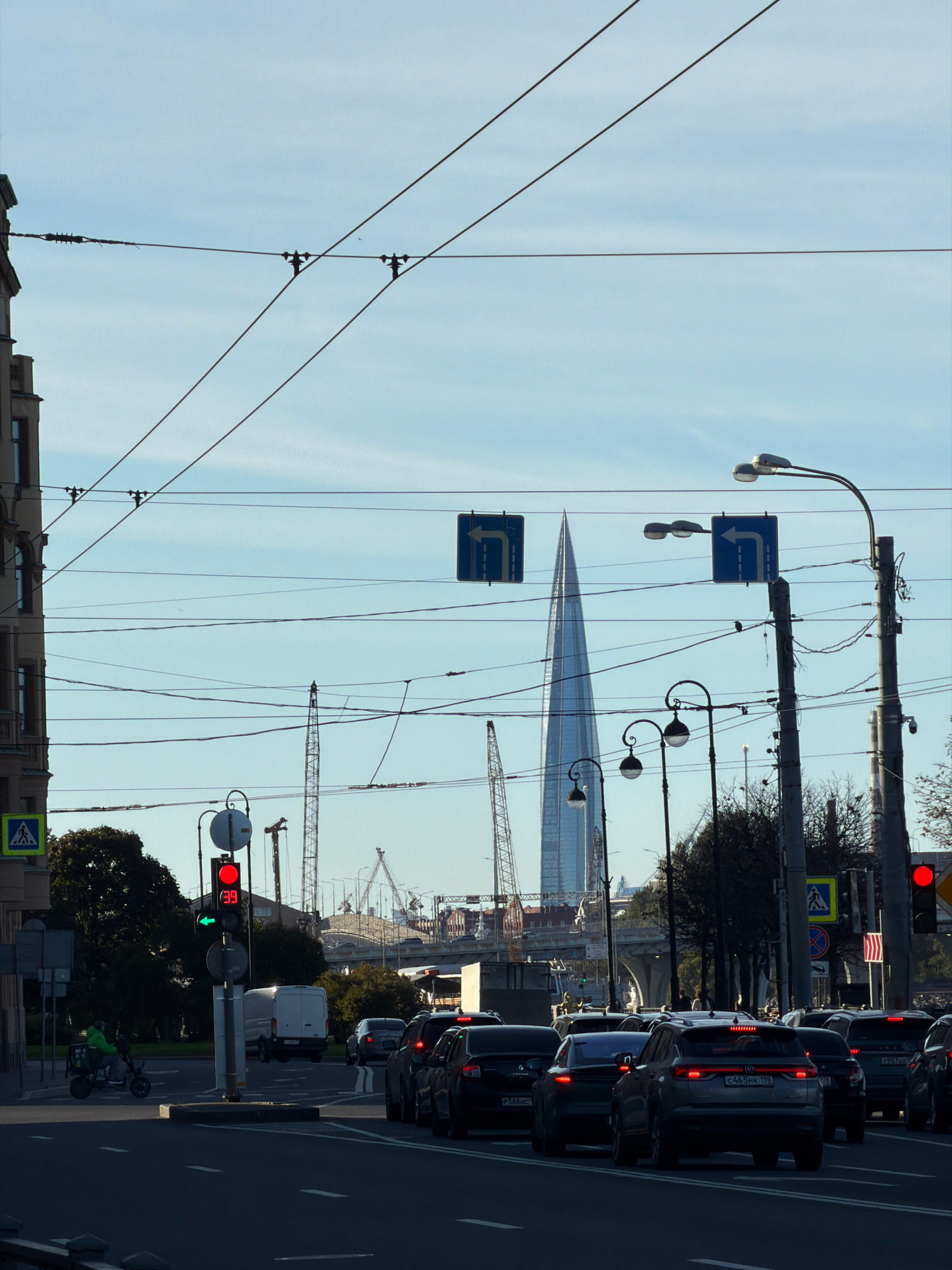 The image depicts a street scene in Saint Petersburg, Russia, with a clear sky and a few clouds. In the foreground, a traffic light shows a red light and a green arrow pointing left. A person on an electric scooter with a green jacket is moving through the intersection. The street is filled with cars, most of which are stopped at the red light. Several traffic signs are visible, including a pedestrian crossing sign and directional signs. In the background, a distinctive tall, glass skyscraper dominates the skyline, identified as the Lakhta Center. Construction cranes are also visible around the Lakhta Center, indicating ongoing development. Overhead, a complex network of power lines crisscrosses the sky, typical of urban infrastructure. The overall scene suggests a busy urban environment during daylight hours.
