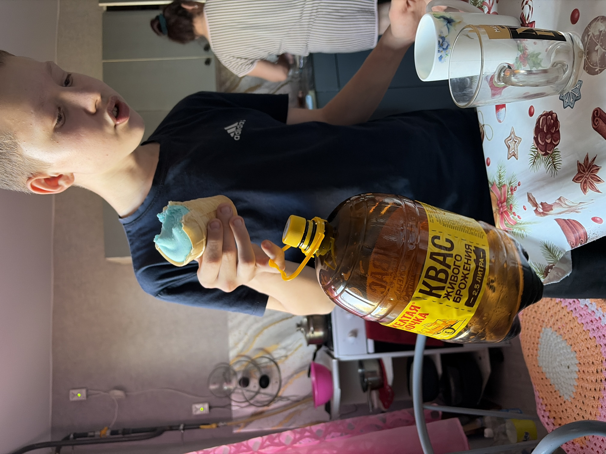 A young person, likely a child, wearing a dark blue Adidas shirt, holds a blue ice cream cone in one hand and a large plastic bottle labeled "KBAC" (Kvas), a fermented Russian beverage, in the other. The label includes the phrase "живого брожения" ("live fermentation"). The person is standing indoors. Behind them, another individual wearing a striped shirt is visible. A table covered with a Christmas-themed tablecloth holding one mug and one glass is in the background. The overall setting appears to be a kitchen or dining area. It is unknown whether this image was taken in the city of Чумна, Russia, and there is no evidence to support the claim that the image was taken there.