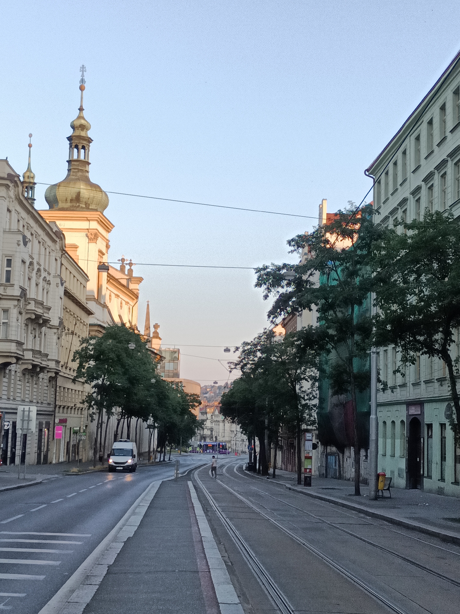 This is a street scene in Prague, Czechia, captured from a low angle looking down a street lined with buildings. The street has tram tracks running down the center. On the left side of the street, there are several historic buildings with ornate facades and a prominent church tower with a golden dome. A white van is driving down the street on the left side. On the right side of the street, there are more buildings and trees. Further down the street, more buildings and hills are visible in the distance. The sky is clear and light blue. A single person is walking on the sidewalk on the right side of the street.