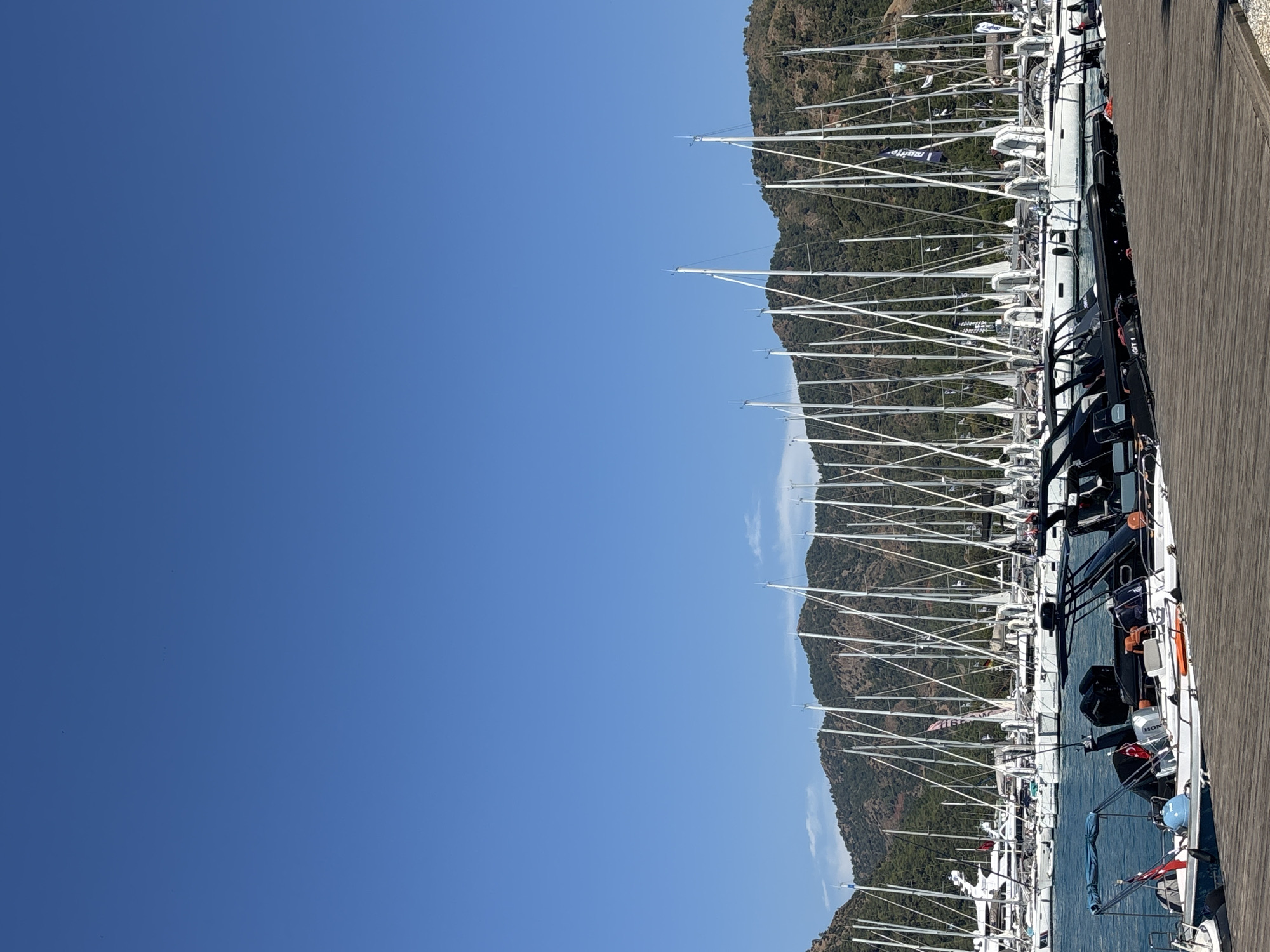 The image captures a serene day at a harbor in Fethiye, Turkey, with clear blue skies dominating the upper half of the frame. The lower portion is filled with a dense array of sailboats, their tall white masts forming a striking visual pattern against the backdrop of lush green, tree-covered mountains. In the foreground, a wooden pier juts into the calm, blue water. Several smaller boats, including a white speedboat with twin outboard motors and a black rigid inflatable boat, are moored along the pier. The speedboat has a folded blue canopy and features a Turkish flag attached to its stern. The overall scene conveys a sense of peace and tranquility, characteristic of a Mediterranean coastal town.
