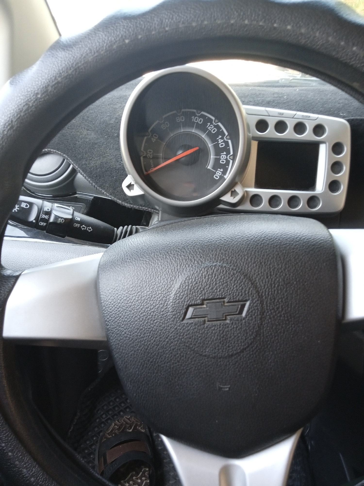 The image shows a close-up view of the interior of a car, focusing on the steering wheel, dashboard, and a portion of the driver's foot. The steering wheel is black and textured, with a silver Chevrolet logo in the center. To the left of the steering wheel is a speedometer with a red needle pointing to approximately 60 km/h. To the right of the speedometer is a silver panel with several circular buttons and a small screen. A black turn signal lever is visible on the steering column below the speedometer. The driver's left foot, wearing a sandal, is visible on the floor mat in the lower left corner of the image. The overall impression is that of a modern car interior.