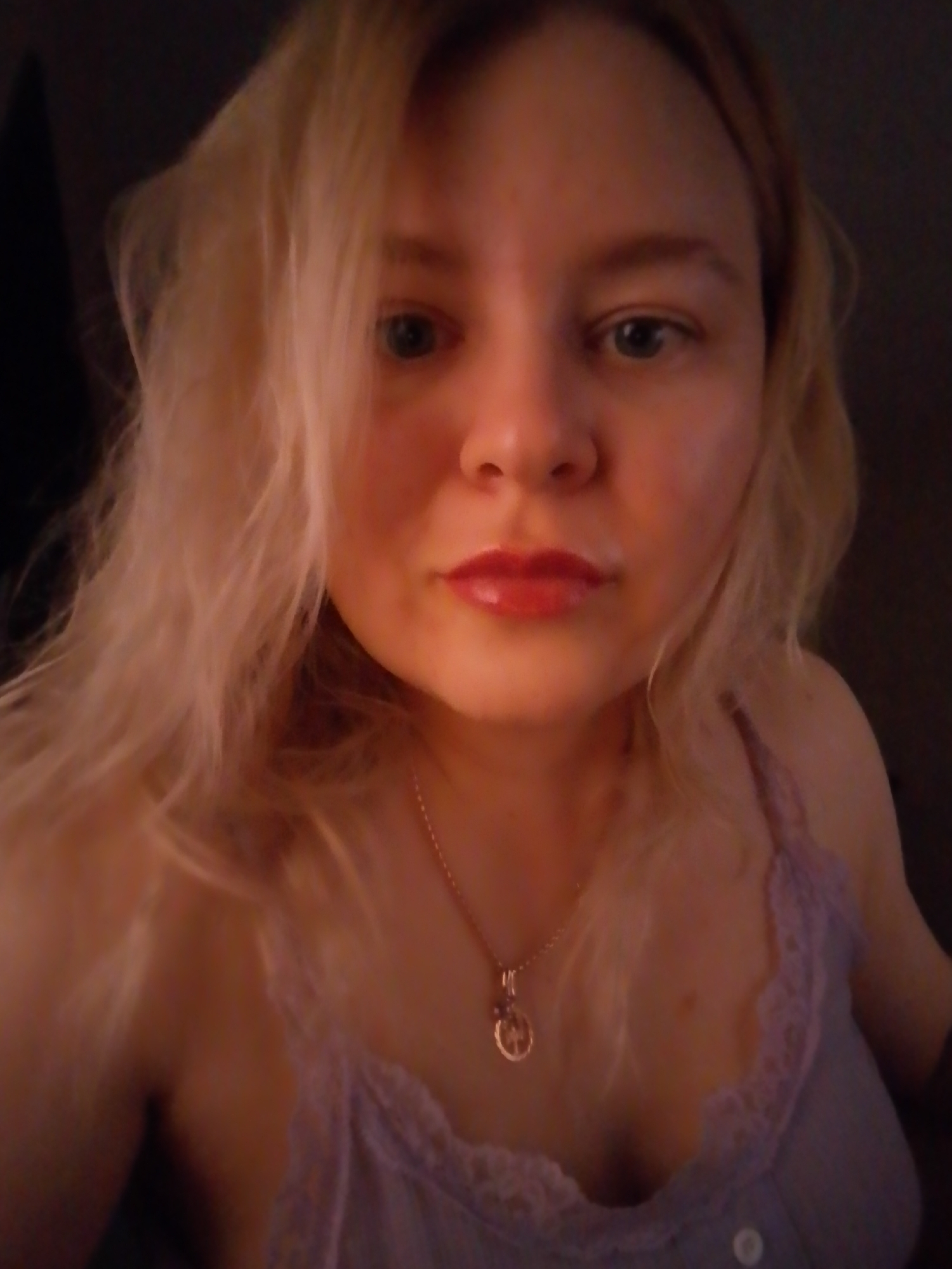 A fair-skinned person with blond hair and blue eyes is posing for a close-up selfie. They are wearing a lavender-colored camisole with lace trim and a gold necklace. The background is dark and out of focus. There is no indication of the location being Izhevsk, Russia.