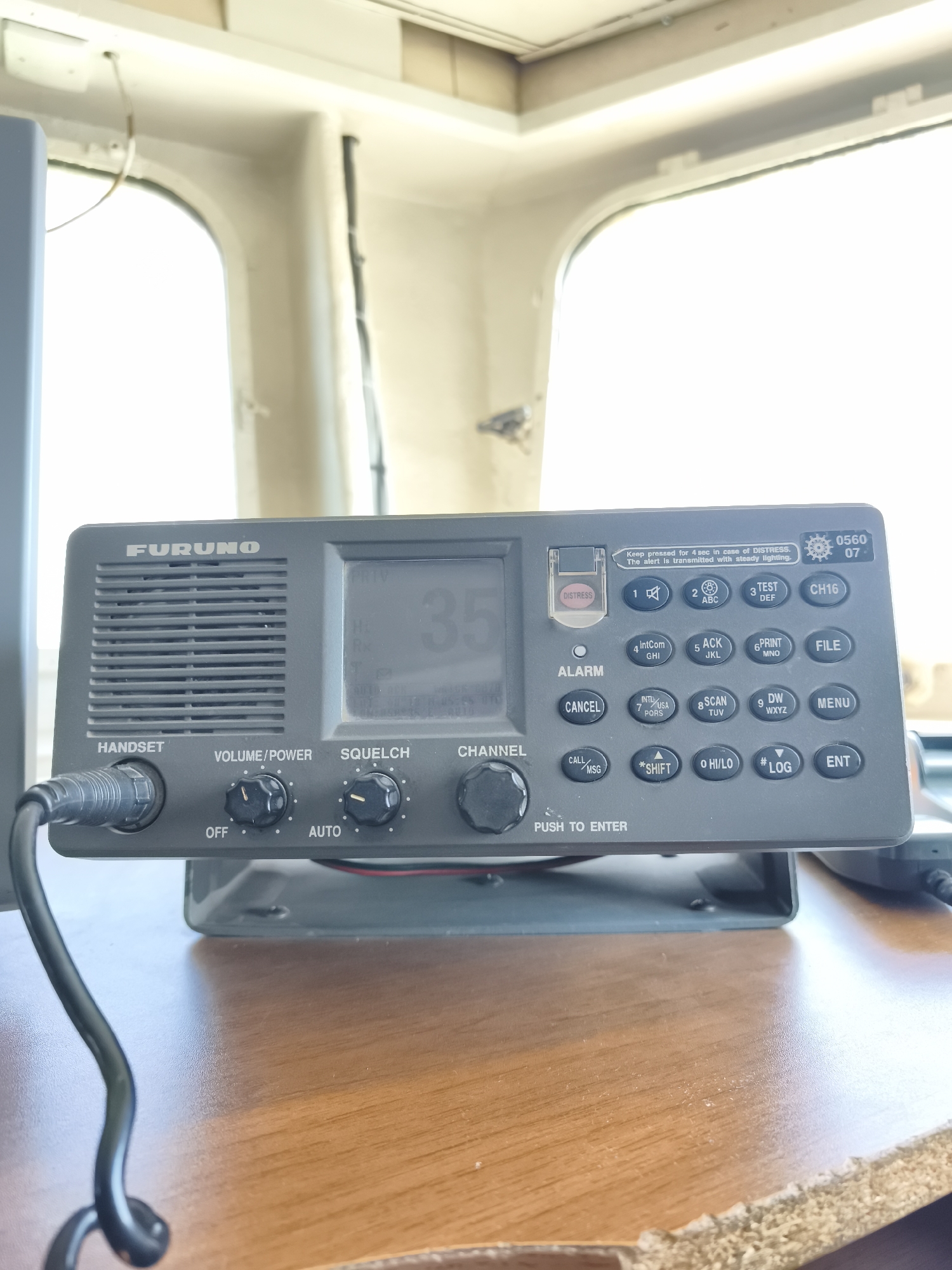 The image shows a close-up of a grey Furuno marine radio located on a wooden surface. The radio has a speaker grill on the left, a digital display in the center showing "35", and a keypad with various buttons on the right. The buttons include numerical keys, "ALARM", "CANCEL", "CALL/MSG", "*SHIFT", "0 HI/LO", "#LOG", "ENT", "INT'L", "PORS", "SCAN", "DW", "WX", "YZ", "FILE", and "MENU". There is also a red "DISTRESS" button with a protective cover. Knobs for "VOLUME/POWER" and "SQUELCH" are visible below the display. A black handset is connected to the radio on the left. The background appears to be the interior of a room with a window, possibly on a boat or in a marine-related building, with bright sunlight coming through the window. The location is identified as Pirallahi Raion, Azerbaijan.