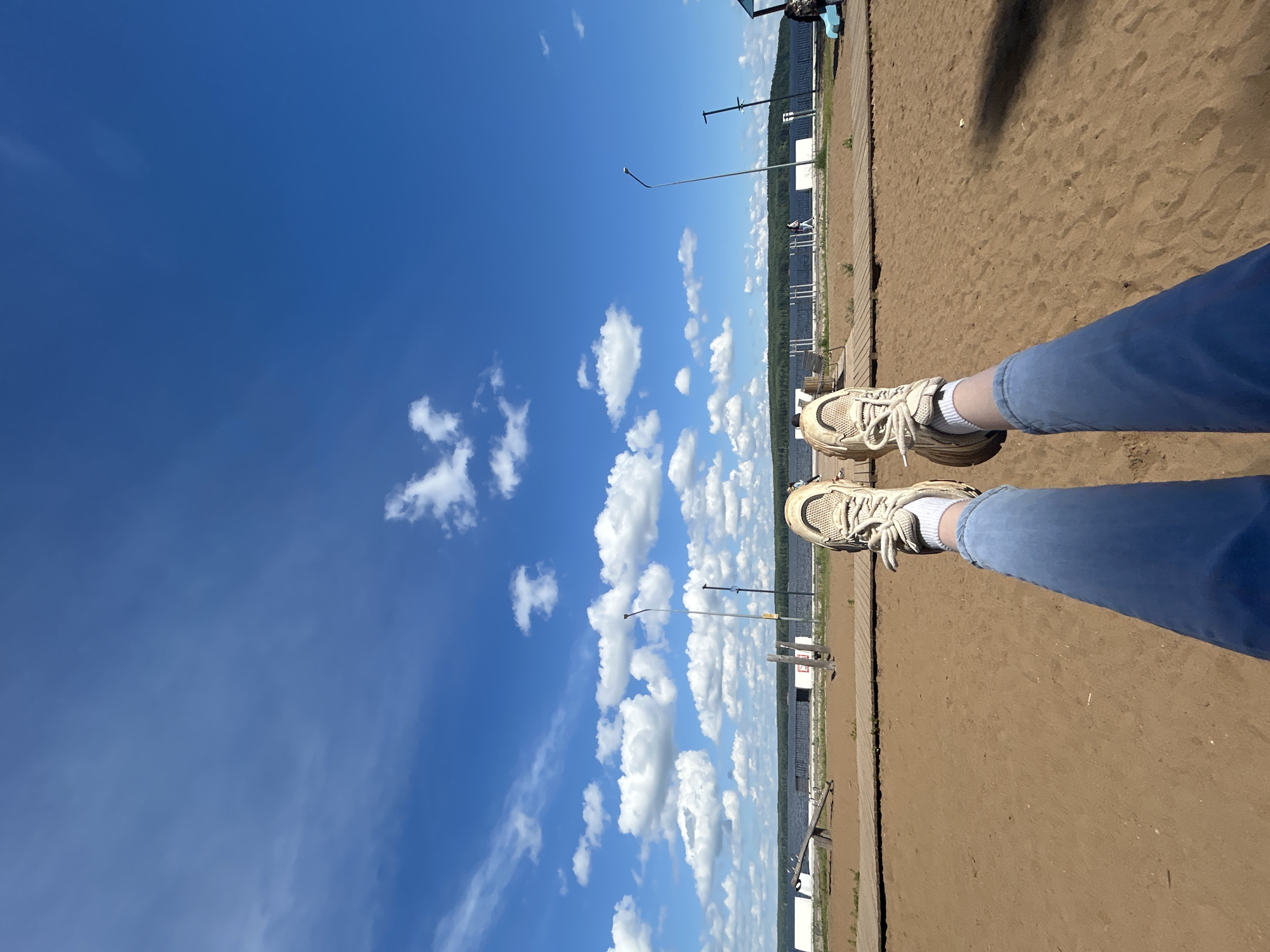 This is a first-person perspective, eye-level outdoor photograph taken on a sunny day with a clear blue sky and scattered white clouds. The foreground is dominated by a sandy beach with a wooden boardwalk running horizontally across the middle ground. The subject's legs, clad in blue jeans and white socks with beige sneakers, are prominently displayed, stretched out towards the camera.
In the middle ground, beyond the beach and boardwalk, is a body of water, likely a river or lake, with a distant treeline visible on the opposite bank. Several lampposts are positioned along the water's edge, and a playground structure, resembling a seesaw, is visible on the left. A few other individuals can be seen walking or standing in the background along the waterfront.
The location is identified as Chaikovsky, Russia, specifically the embankment area. The overall scene depicts a recreational setting, likely a park or beach area along the Kama River.