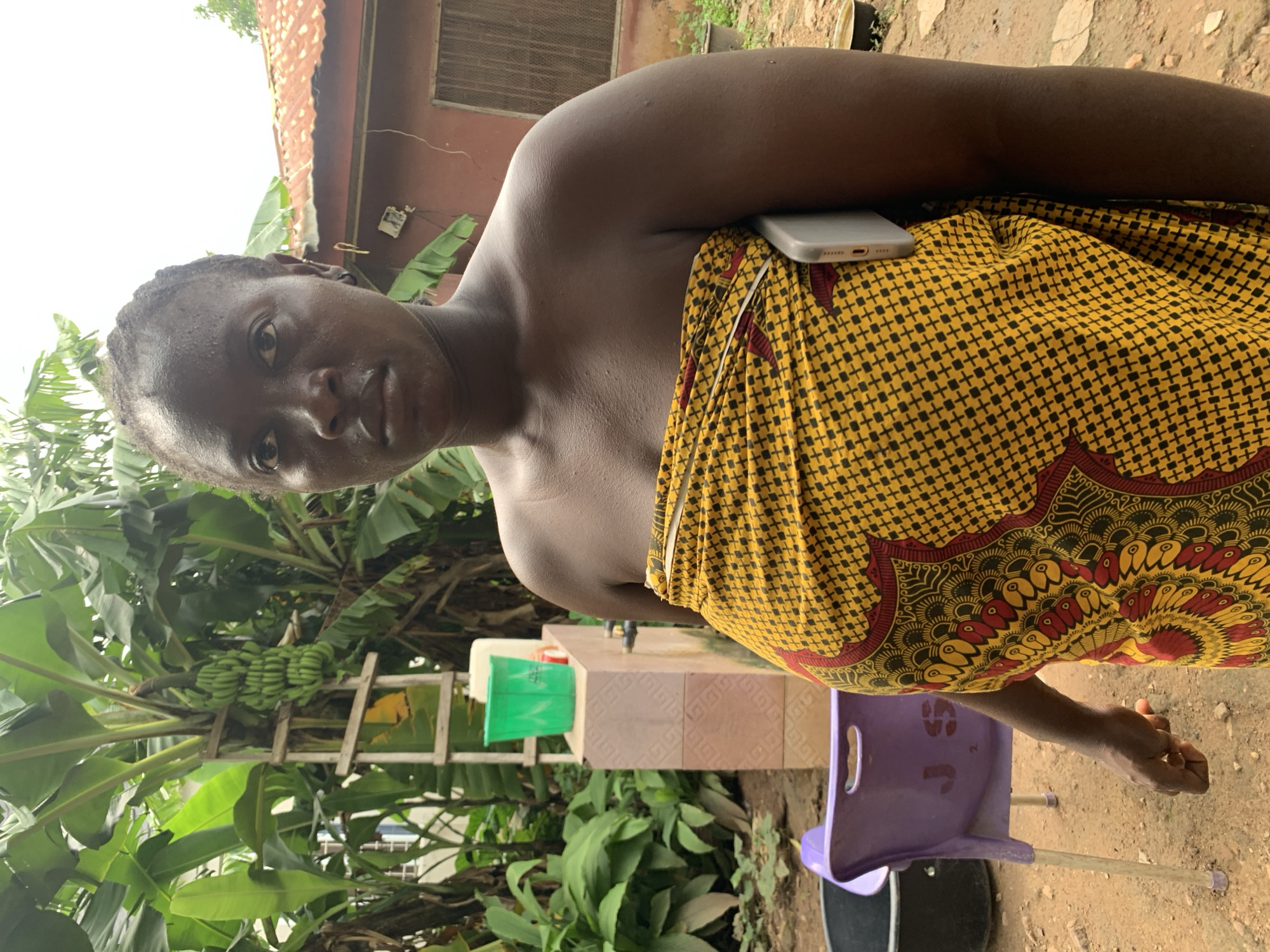 A dark-skinned woman with braided hair stands in front of a building, wearing a yellow and black patterned cloth wrapped around her body. A gray smartphone is tucked into the cloth on her right side. Behind her, banana trees with bunches of green bananas are visible. To the left of the woman, a purple chair is overturned on the ground, and further back, a green bucket sits atop a tiled structure. The ground is sandy and uneven, with some scattered debris. The building behind her appears to be made of brick with a corrugated metal roof. The scene suggests an outdoor, possibly rural or semi-urban, setting. The specific location is Akwanga, Nigeria.