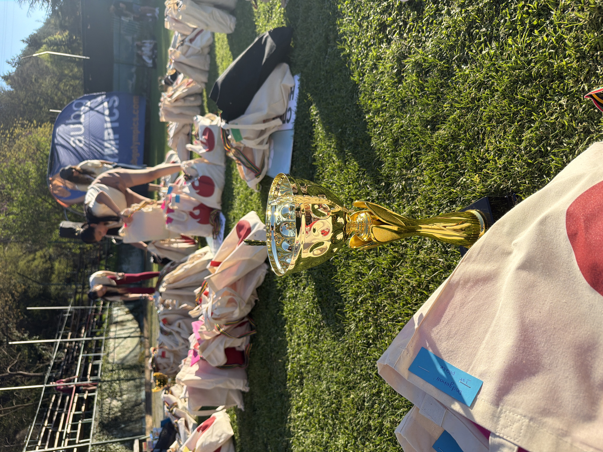 The image shows a gold-colored trophy on top of a pile of bags in a grassy area. A blue tag on one of the bags reads "Dodgeball, 1st place." In the background, people are interacting near a blue banner with "aubg Olympics" printed on it. The location is likely in or near Blagoevgrad, Bulgaria, given the "aubg" banner, which stands for American University in Bulgaria, located in Blagoevgrad. The scene appears to be an outdoor award ceremony or gathering after a dodgeball event.