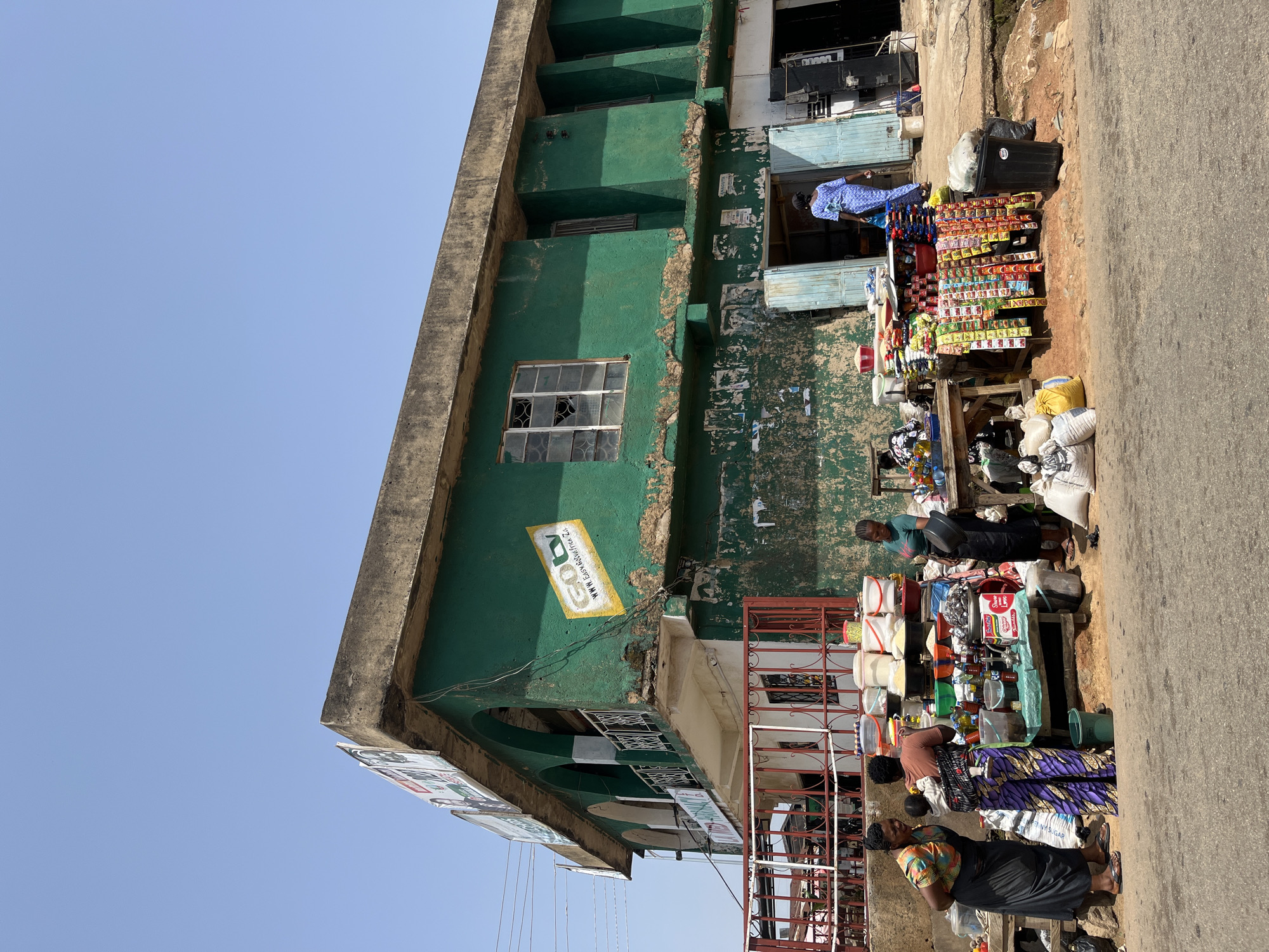 The image depicts a street scene in Jos, Nigeria, under a clear, bright sky, suggesting midday. A weathered, green building with a concrete facade dominates the left side of the frame. Its walls are peeling and stained, with a broken window visible on the first floor. A yellow sign with black text, reading "GO" and "www.LEGYMASSAGEFREE.CZ", is affixed to the green wall.
In front of the building, a bustling marketplace is set up along a paved road. Several people, primarily women, are engaged in commerce. They are standing around stalls laden with various goods, including sacks of what appears to be flour or sugar, buckets, bottled drinks, and packaged food items. One woman, wearing a blue and white patterned garment, is seen standing near a stall. Another woman in a purple and black patterned dress is interacting with a vendor. Further down, a woman in a colorful patterned top and black skirt is also present.
The scene suggests a typical day in a Nigerian market, with vendors selling their wares and customers browsing and purchasing. The overall atmosphere appears active and industrious, characteristic of a busy market environment. The sunlight casting shadows indicates it is daytime.