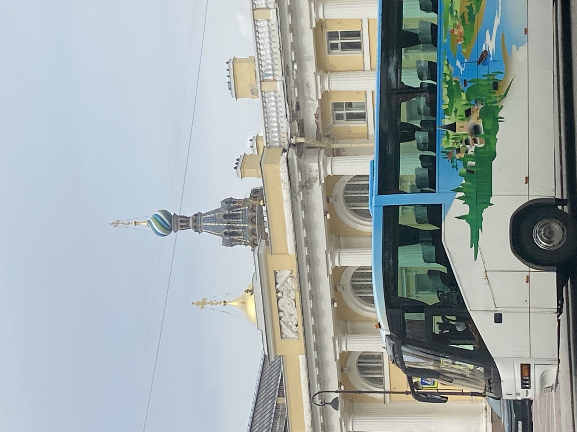 A tour bus with a colorful mural on its side is parked in front of a pale yellow building with classical architecture. The building features a colonnade with Ionic columns and arched windows. On top of the building, a golden dome with a cross is visible, along with a blue and green striped onion dome of a church. The bus is white with a blue upper section and a mural depicting a town with a church, river, and trees. The sky is overcast. The scene appears to be in Saint Petersburg, Russia, given the architectural style and the presence of distinct onion domes.