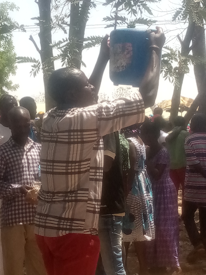 In Kagoma, Nigeria, an outdoor scene unfolds under bright daylight. A person with dark skin, wearing a light-colored plaid long-sleeved shirt and red trousers, stands prominently in the foreground, facing right but looking upwards. This individual holds a blue plastic jerrycan, approximately 5-10 liters in capacity, above their head with both hands, appearing to either pour liquid or access its contents. The top of the person's head and parts of their upper body appear wet, consistent with water or another liquid. In the blurred background, a crowd of approximately 10-15 individuals, including adults and children dressed in various casual attire, such as plaid shirts and patterned dresses, are visible. The environment features sparse trees with light green foliage and an unpaved earthen ground. A light-colored, possibly temporary, structure or tent is discernible in the far right background.