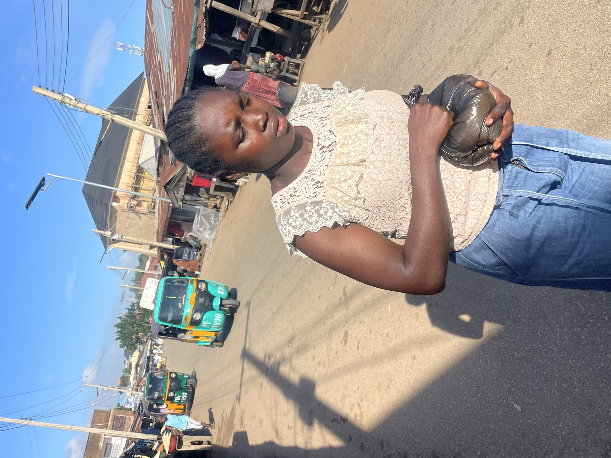 In Jos, Nigeria, a young African woman with braided hair walks down a street lined with shops. She wears a light pink lace top and blue jeans, and carries a black plastic bag in her hands. The street is paved, and there are several tuk-tuks, a common mode of transport in the area, on the road. The buildings in the background appear to be a mix of residential and commercial structures, with some showing signs of wear and tear. The sky is clear and blue, indicating a sunny day. The overall scene depicts a typical day in a bustling Nigerian city.