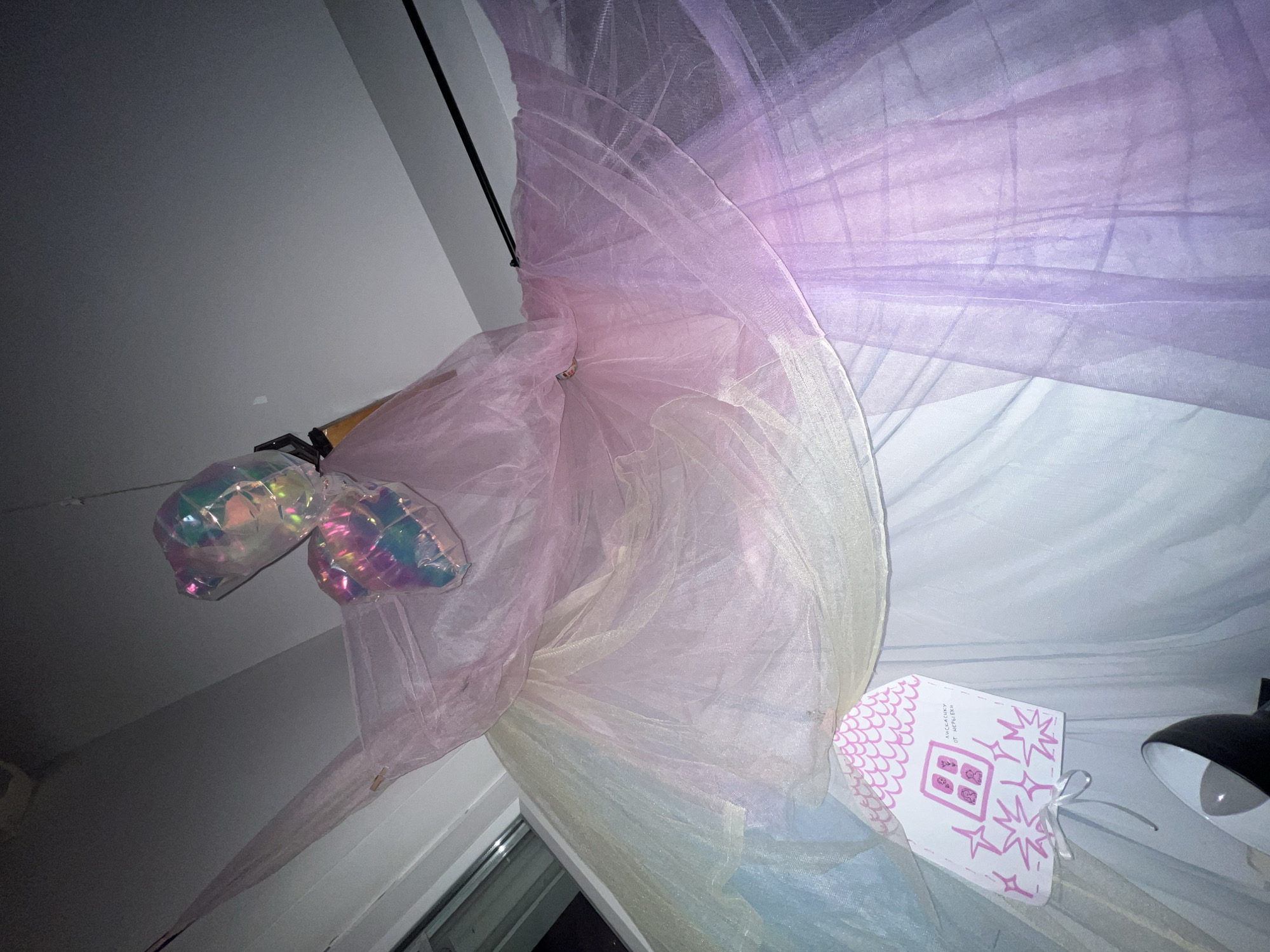 A low-angle indoor shot shows a window adorned with pink and purple sheer fabric. Two iridescent balloon-like objects hang near the window. To the right, a white gift bag with pink designs and a tied ribbon is present. A black lamp is visible in the lower right corner of the frame. The setting seems to be in an interior space, potentially a bedroom or living area. The location is Palilula Urban Municipality, Serbia.
