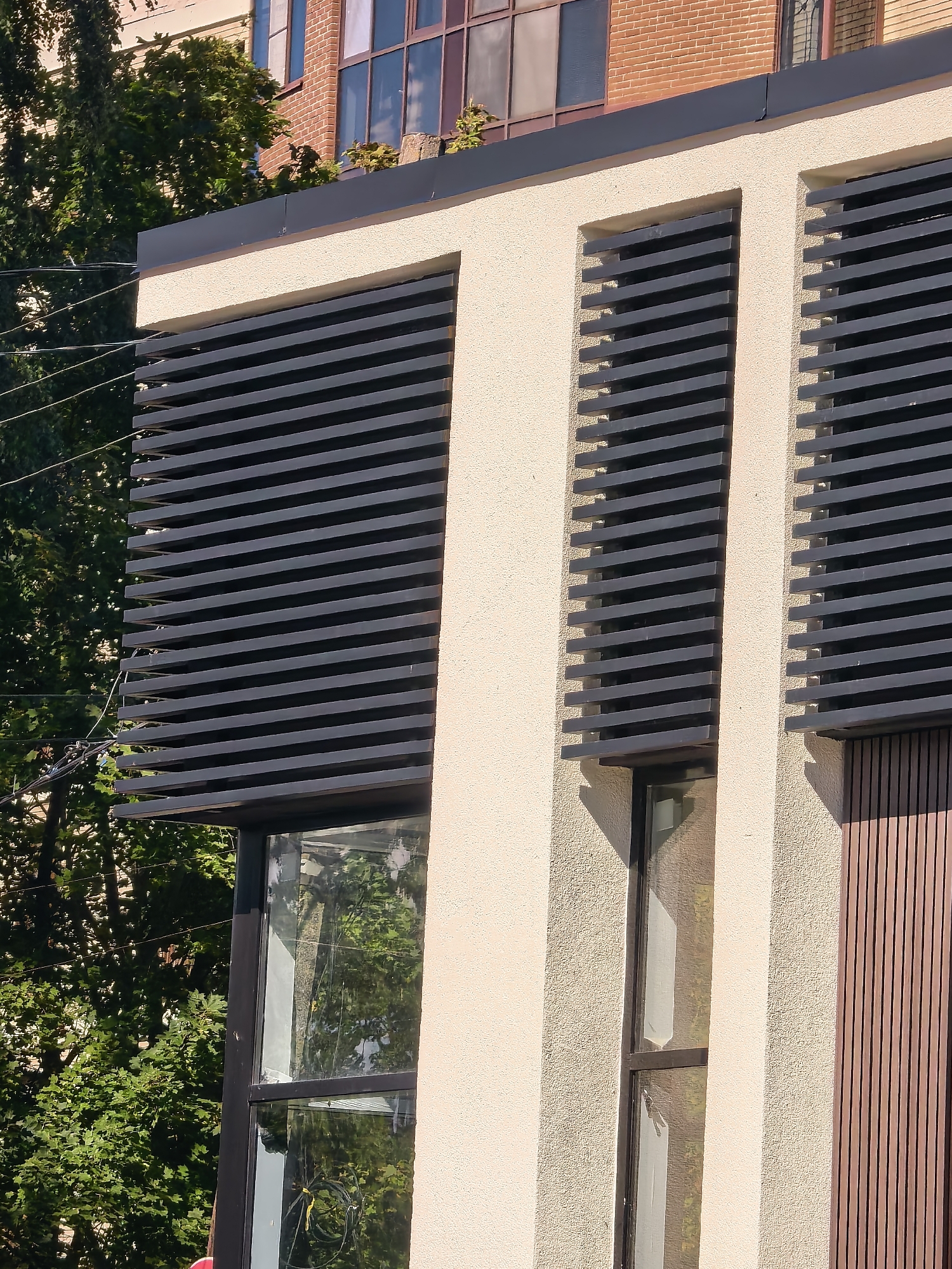 The image shows a close-up of a modern building exterior in Sestroretsk, Russia. The building has a textured, light-colored stucco facade. Prominently featured are horizontal dark gray louvers covering sections of the building, appearing to be part of a fenestration system. Below the louvers, dark-framed windows are visible, reflecting greenery and sky. To the right of the louvers, a section of the facade is clad with vertical wooden planks. In the background, partially obscured by the modern building, is an older brick building with multi-paned windows. Lush green trees and some power lines are also visible in the background. The lighting suggests it is daytime, with sunlight casting shadows. There are no people or animals in the frame.