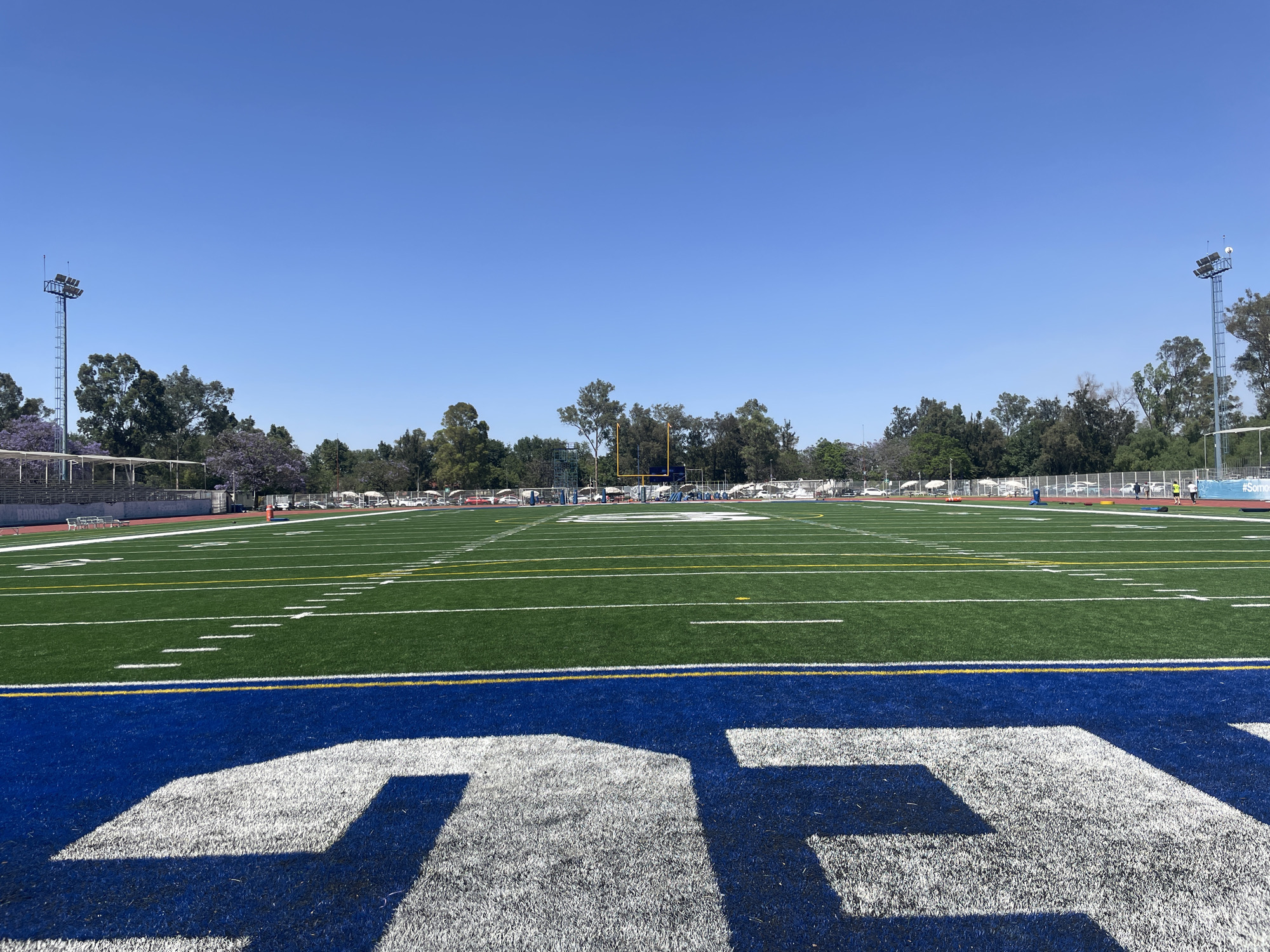 This image displays an American football field under a clear, bright blue sky in Mexico City, Mexico. In the foreground, the endzone is painted a vibrant blue, featuring large, stylized white letters partially visible, suggesting part of an abbreviation like "CDCC" or "GDC". The field itself is artificial green turf, precisely marked with white and yellow lines indicating yardage and boundaries. A red running track encircles the entire playing area.
A yellow goalpost is visible at the far end of the field. The background is lined with lush green trees, some distinguished by their purple flowers, characteristic of Jacaranda trees. Tall stadium lights with multiple lamps are positioned on both the left and right sides of the field. To the left, bleachers are visible, with the words "JACARANDAS" and "BORREGOS" painted on a wall or banner. On the far right, a blue banner displays the hashtag "#Somos". Beyond the field's fence, some vehicles and a few individuals are faintly visible walking. Strong shadows cast across the field suggest a sunny midday or early afternoon. The scene is calm and appears prepared for athletic events.