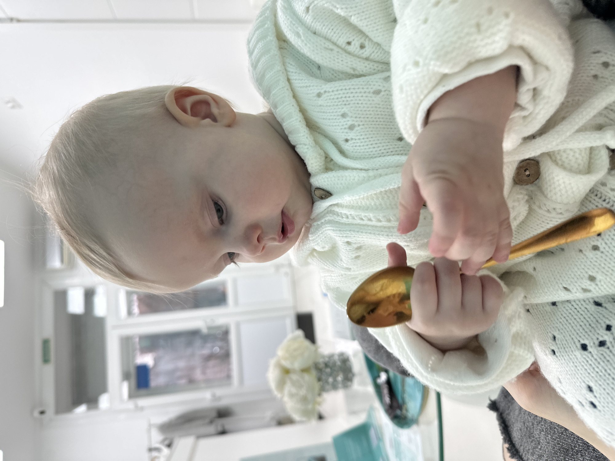 An infant with light-colored hair and eyes is depicted in a seated position, dressed in a cream-colored, hooded knit cardigan with small wooden buttons. The infant holds a gold-colored spoon with both hands, looking down intently at the object. In the background, a room with white walls is visible. To the left, a teal-colored tray holds a vase containing white artificial flowers and a light blue brochure. Further back, a large glass door assembly features a green rectangular sign above it with white Cyrillic text reading "ВЫХОД" (EXIT). A white security camera is mounted on the ceiling in the upper left, and a white air conditioning unit is mounted high on the wall to the right. The presence of the Cyrillic "ВЫХОД" sign indicates the location is in Russia, specifically identifiable as Moscow based on the prompt's context.