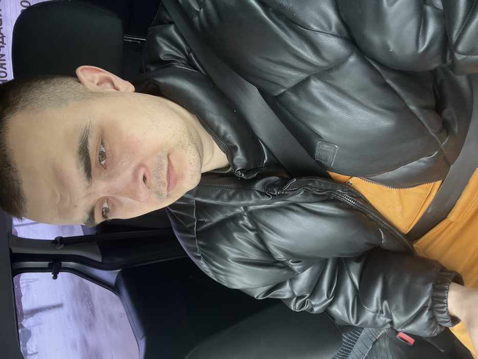 A male individual is seated in the front passenger seat of a vehicle in Novosinkovo, Russia. He has short, dark hair, light facial stubble, and light brown eyes, with his gaze directed downwards and to the right. He wears a black, quilted, synthetic leather puffer jacket, partially zipped, over an orange garment. A black seatbelt is buckled across his chest. The vehicle's interior is dark, with a patterned fabric visible on the seat to the subject's left. Through the blurred side window, a cold, possibly snowy or overcast outdoor environment is visible, characterized by indistinct white and grey tones and faint linear elements. Partial Cyrillic text, including 'КОНЧД', is faintly discernible on a white background in the upper left of the exterior view.