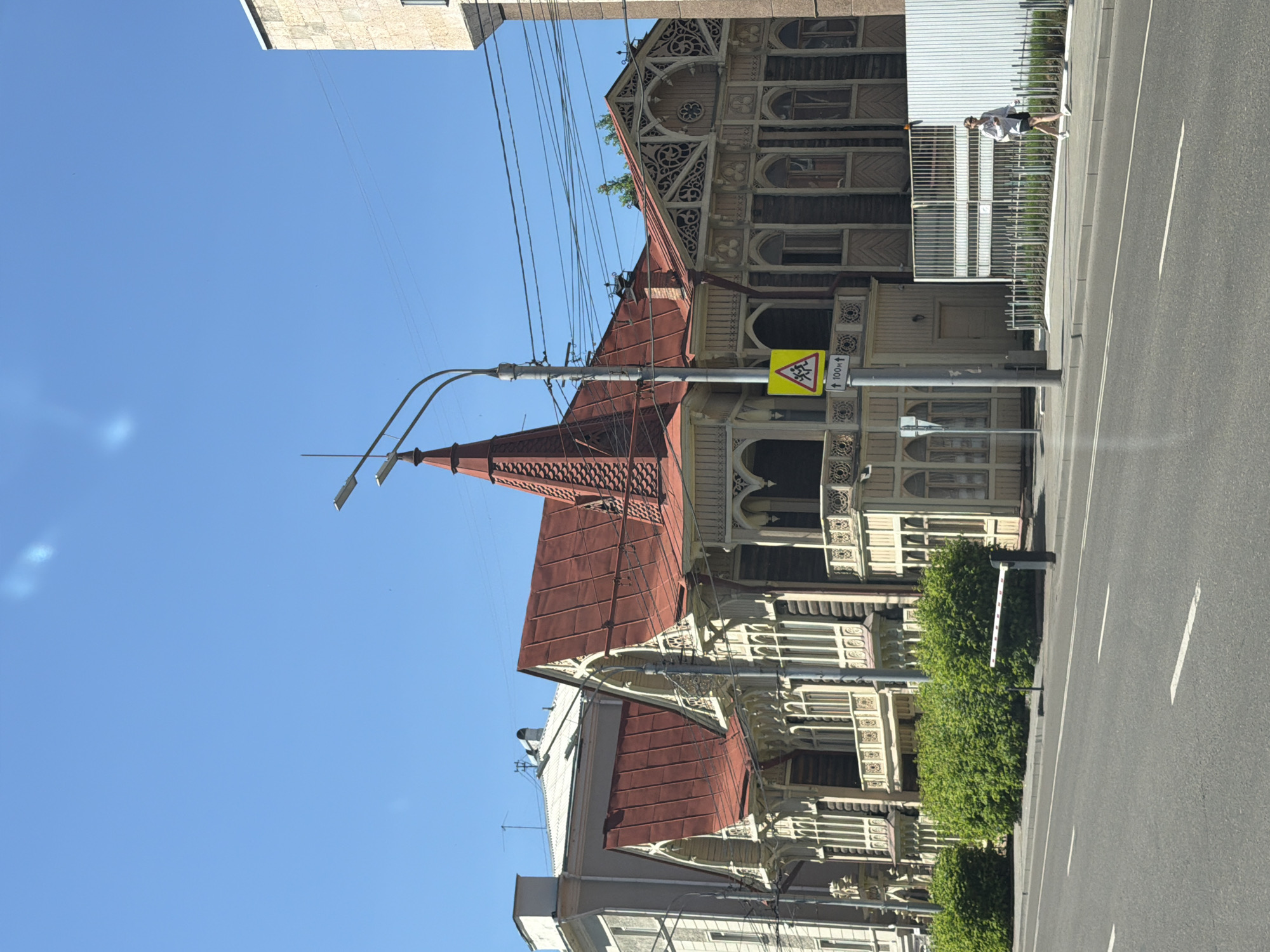 This image depicts a street scene in Krasnoyarsk, Russia, on a clear, sunny day. The main subject is a historic wooden building with intricate carvings and a red, multi-tiered roof, characteristic of traditional Russian architecture. The building has multiple windows and balconies, adorned with decorative wooden elements.
In the foreground, a paved road with white lane markings runs across the bottom of the frame. A sidewalk runs alongside the road, separated by a low metal fence and green bushes. A white barrier arm is visible across the sidewalk, suggesting a controlled entrance or passage.
On the sidewalk, a young woman is walking to the right. She is wearing a white top, dark shorts, sunglasses, and white sneakers. She is carrying a dark bag over her right shoulder.
A street light with multiple bulbs stands on the sidewalk, and a yellow traffic sign with a silhouette of children and an arrow indicating "100m" is mounted on the same pole. Power lines are strung across the sky, typical of urban environments.
Behind the wooden building, other structures are visible, including a modern building to the far right and older buildings with white and grey facades further to the left. The sky is a bright, cloudless blue. The overall impression is a snapshot of an urban environment with a blend of historical and modern elements.