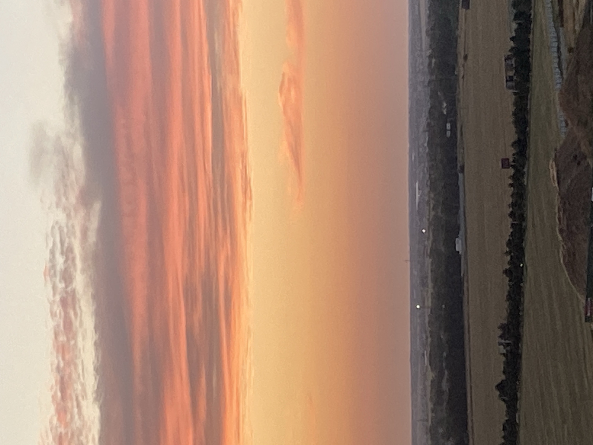 This is an aerial, close-up, eye-level, and medium shot image of a landscape during sunset. The sky is filled with vivid orange and pink hues from the setting sun, with horizontal bands of clouds stretching across the frame. Below the sky, a distant horizon shows a flat landscape with scattered trees, fields, and some structures. The foreground is out of focus and shows a brown, rough surface, possibly earth or construction material. The image is located in Chornomorsk, Ukraine.