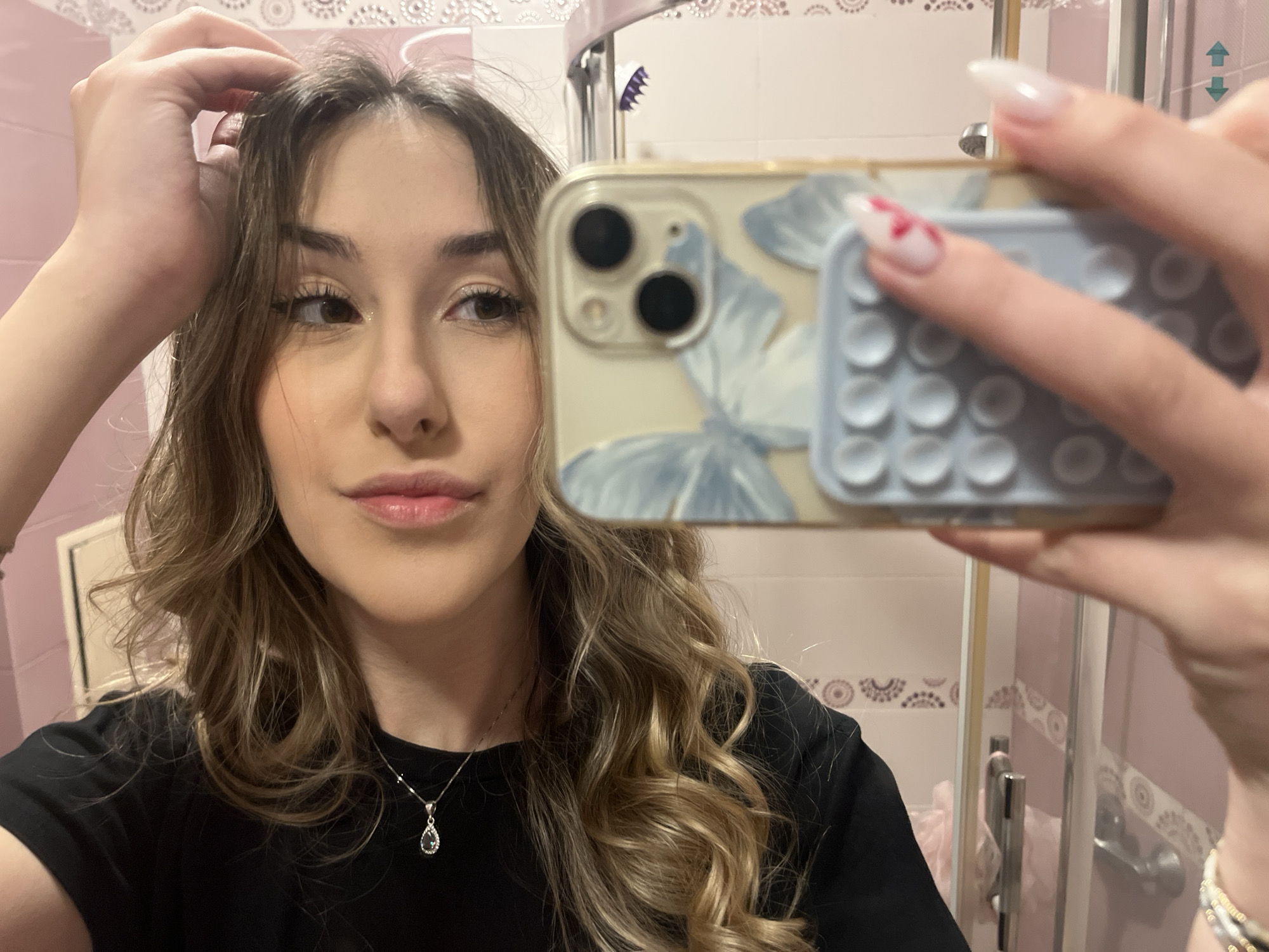 A young woman is taking a selfie in a bathroom. The bathroom has pink walls and decorative tile borders. The woman is wearing a black shirt and a necklace with a teardrop-shaped pendant. Her hair is long, wavy, and light brown with blonde highlights. She is holding an iPhone with a butterfly-patterned case. The phone is obscuring part of her face. She has long, white, pointed nails, with one nail painted with a red floral design. The background includes a shower stall with a transparent door and silver fixtures. The location is likely a private residence. There is no information to indicate this is located in Sofia, Bulgaria.