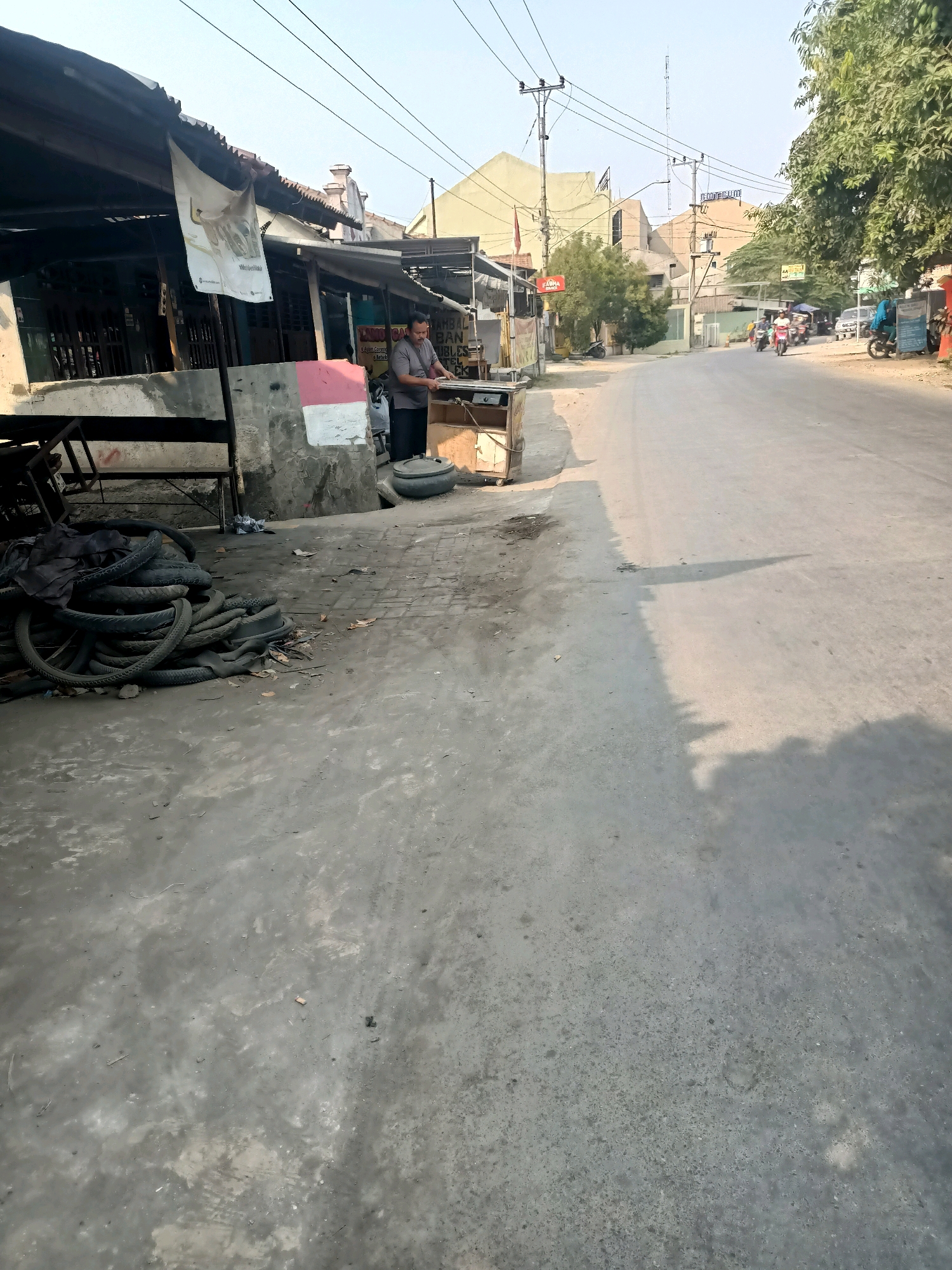 A man stands in front of a stall with a tarp roof on the side of a paved street in Tembok Banjaran, Indonesia. The man is wearing a gray polo shirt and black pants. He is holding a piece of metal. In front of the stall, there is a pile of tires. The street is mostly empty, with a few motorcycles and cars in the distance. Buildings line the other side of the street. The sky is clear and the sun is shining.