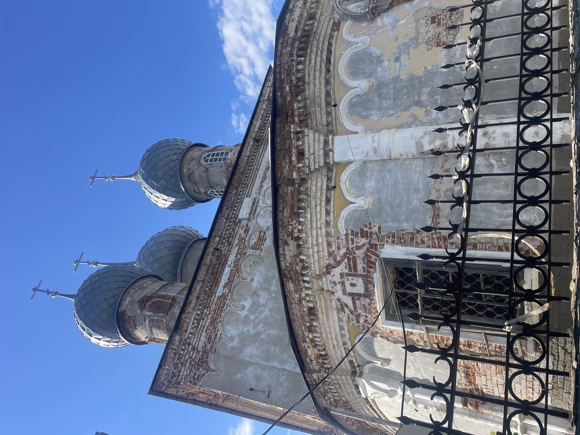 A low-angle shot captures the domes and facade of a weathered brick church under a clear blue sky with scattered clouds. The church features multiple onion domes covered in gray, scale-like shingles, each topped with a cross. The brickwork of the facade is exposed in many areas, showing signs of age and deterioration with peeling plaster revealing the underlying red bricks. Decorative architectural elements, such as arched window frames and cornices, are visible. A black wrought-iron fence with decorative finials and circular motifs runs across the foreground, partially obscuring a window with a metal grate. The scene appears to be in Yuryevets, Russia.