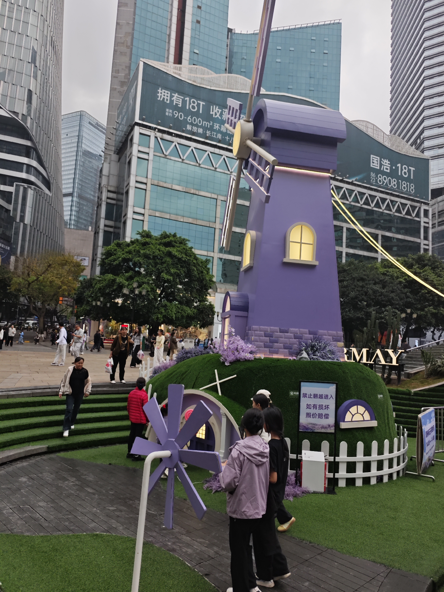 The photograph shows a whimsical purple windmill display in Yuzhong District, China. A large purple windmill with lit windows and faux stonework stands on a green, turf-covered mound. At the base, there is a white picket fence and a sign in Chinese characters that reads, "禁止翻越进入 如有损坏 照价赔偿," which translates to "No climbing, damage will be compensated." Another smaller purple windmill sits in front of the display.
Several people are visible in the scene: some standing in front of the windmill, possibly interacting with the display, and others walking on the plaza. The plaza includes green stepped areas, and the ground is partly covered with synthetic turf.
In the background, tall, modern buildings surround the plaza. Billboards advertise "国浩·18T" with a phone number (8908 1818) and "拥有18T 收" followed by "90-600m²环幕 解放碑 长江岸 十" on one building. "MAY" is spelled out in large letters on the side of the mound. The sky appears overcast.