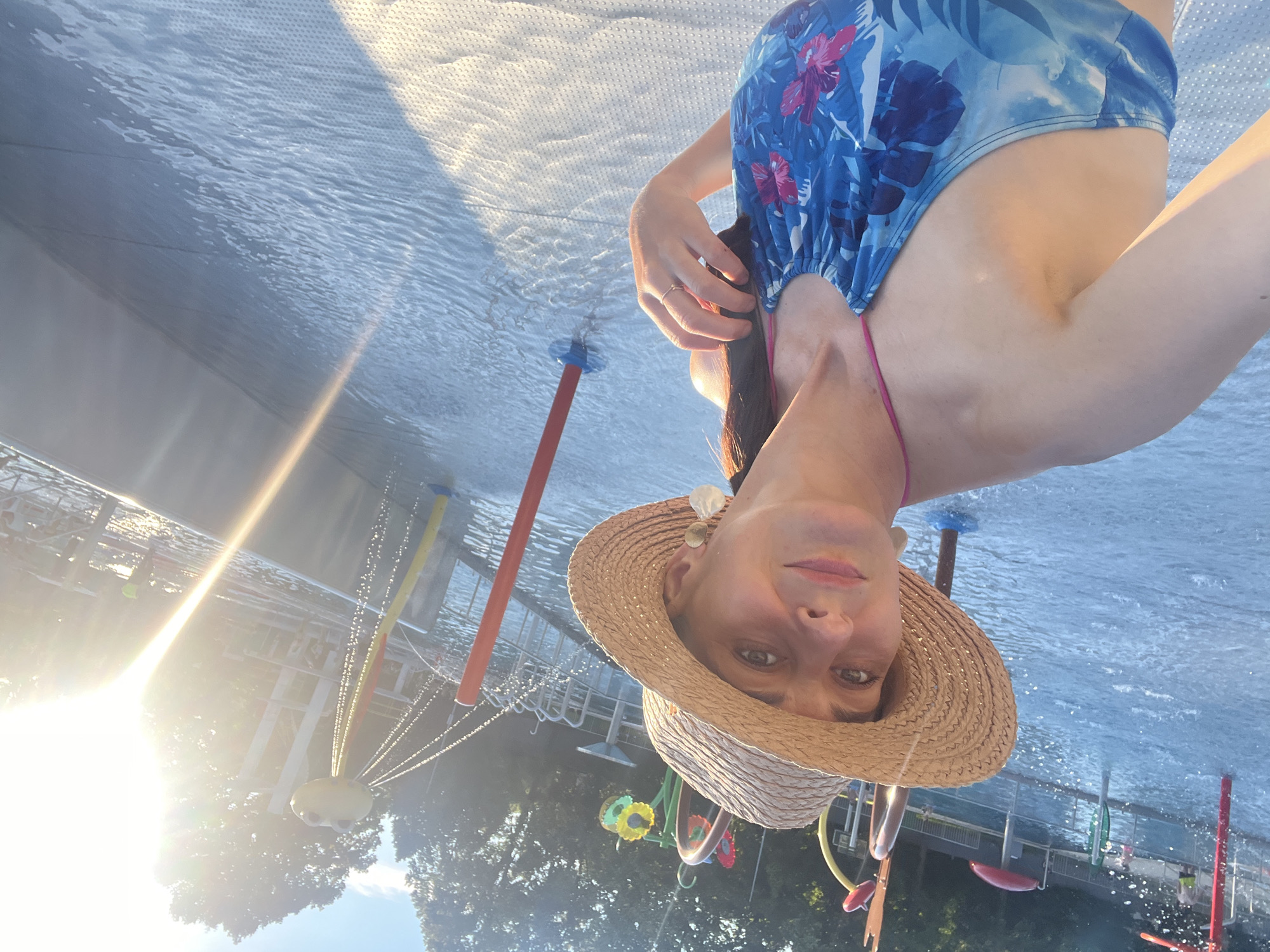 In a bright, sunny outdoor setting, a woman in a blue floral bikini and a straw hat poses for a selfie. She is standing in a shallow water play area with numerous fountains and colorful structures spraying water. The woman is looking towards the camera with a slight smile. The background shows more of the water park with trees and structures visible. The sun is setting, casting a warm glow on the scene.