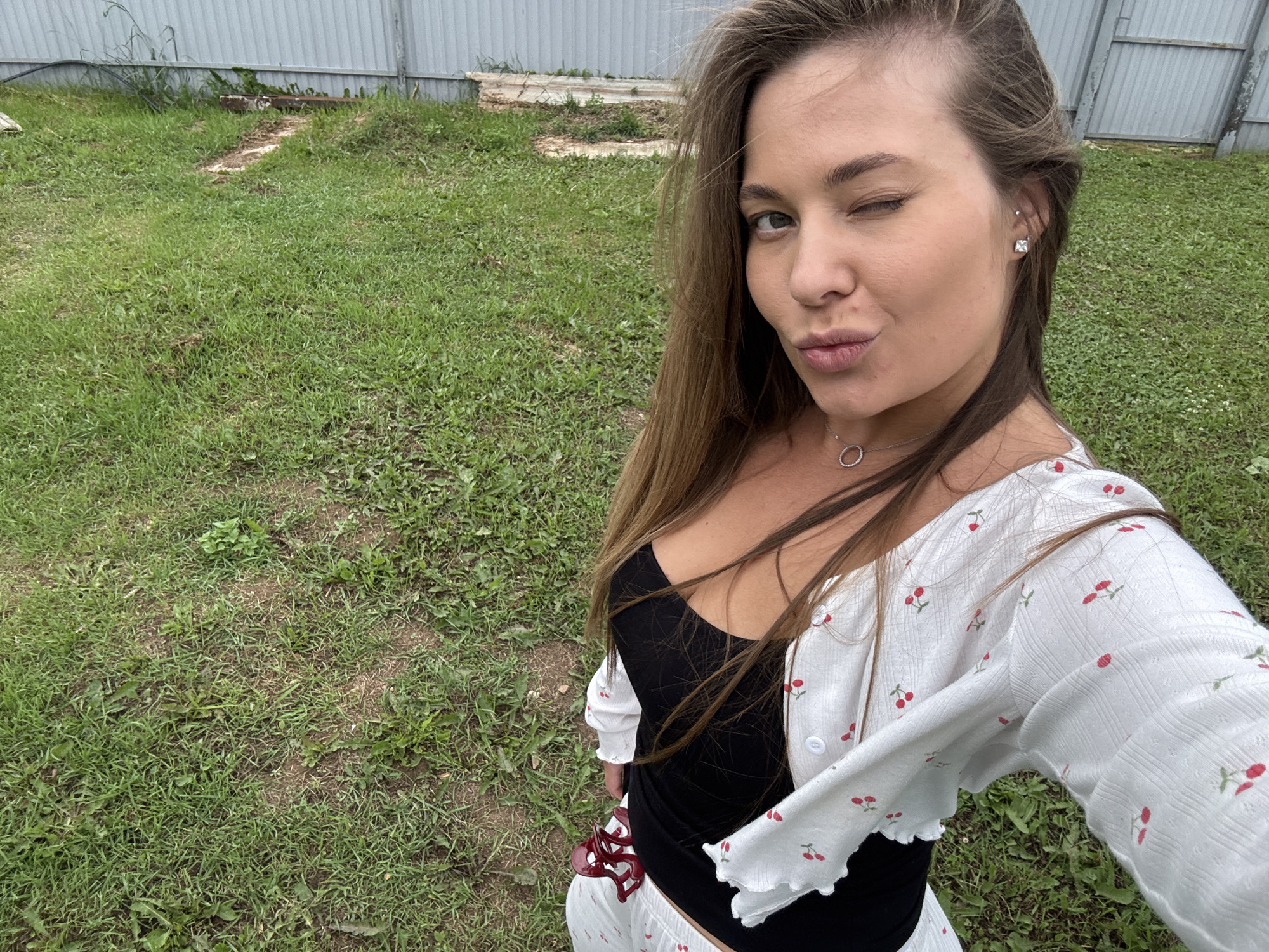 A woman stands in a grassy yard, winking at the camera. She has long, brown hair and is wearing a black top and a white cardigan with a cherry pattern. The background shows a grey metal fence and some overgrown grass. The location is the city of Perm, Priamurye, Russia.