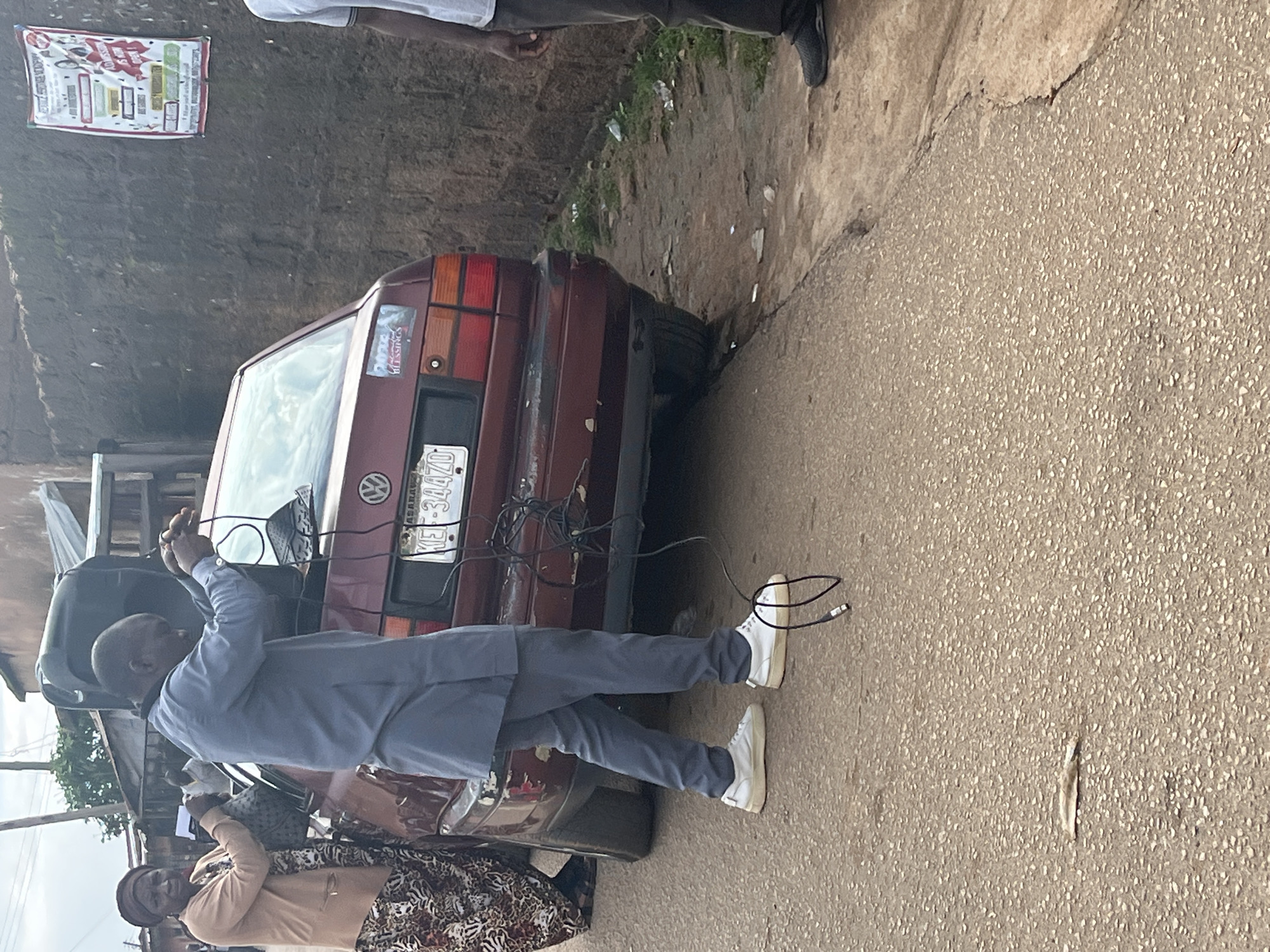 In the city of Jos, Nigeria, a man in a grey traditional outfit and white sneakers stands next to a maroon Volkswagen car, actively connecting cables to a speaker placed on the trunk. To the left, a woman wearing a headscarf and a patterned skirt observes the scene. The car's rear license plate is visible, reading "KEI 34470". The environment suggests a street with a rough asphalt surface and a textured brick wall in the background, adorned with a poster. Other individuals are present in the background, and the scene appears to be outdoors under an overcast sky.
