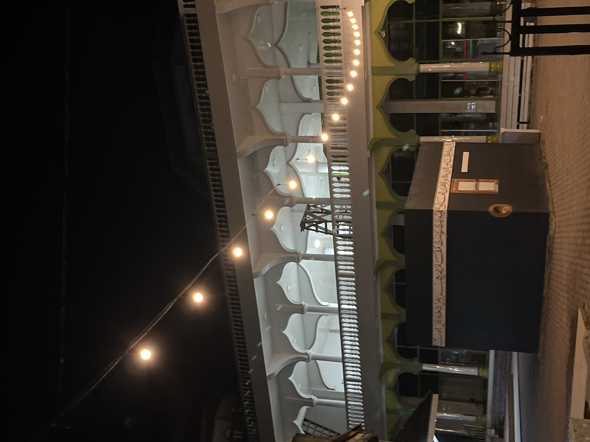 The image depicts the exterior of a mosque at night in Pulodolang, Indonesia. A replica of the Kaaba, a black cube with Arabic calligraphy on its side, is positioned in the foreground. The mosque itself features a white facade with arched windows and a long, white balustrade. String lights are visible overhead, illuminating the scene. The ground is paved with brick. A ladder is leaning against the mosque's structure, suggesting ongoing construction or maintenance. The overall atmosphere is serene and religious, typical of a place of worship at night.