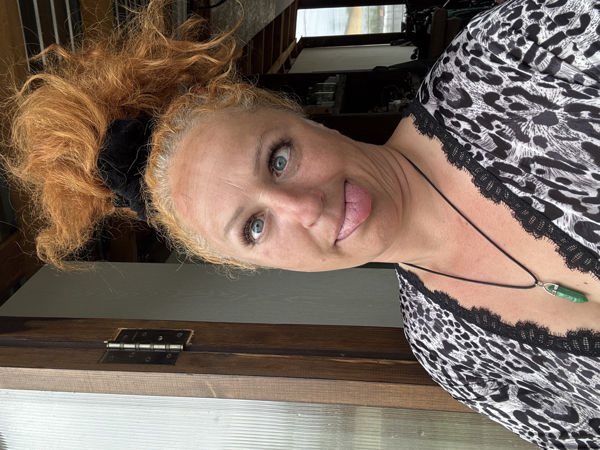 A middle-aged Caucasian woman with bright red, messy hair pulled up in a bun, and a black scrunchie, sticks her tongue out at the camera. She has blue eyes, heavy mascara, and a leopard-print top with black lace trim. She is wearing a black cord necklace with a green, crystalline pendant. The woman is standing outside a building with a wooden door frame, a visible metal door hinge, and a white corrugated plastic panel on the left. The background shows an interior with shelves and some equipment, possibly a workshop or storage area, and a window with a view of a hazy, overcast sky. The location is identified as being in the city of Chaikovsky, Russia.