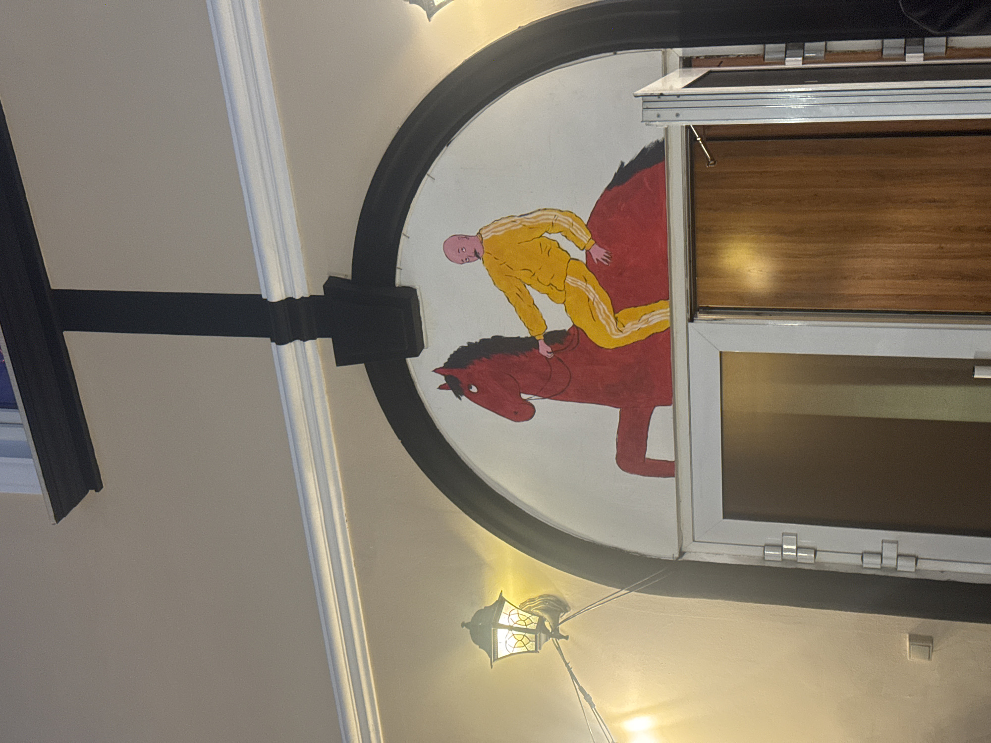 The image depicts a mural of a man in a yellow tracksuit riding a red horse. The mural is painted on a white arched wall above a doorway. The man has a bald head and a thin mustache, and the horse has a long mane and tail. A black structural element, resembling an inverted cross, is situated directly above the mural. To the left of the mural, a vintage-style lantern is mounted on the wall. The doorway below is partially open, revealing a wooden door. The overall scene suggests an interior space with decorative elements.
This image was taken in Krasnoyarsk, Russia.