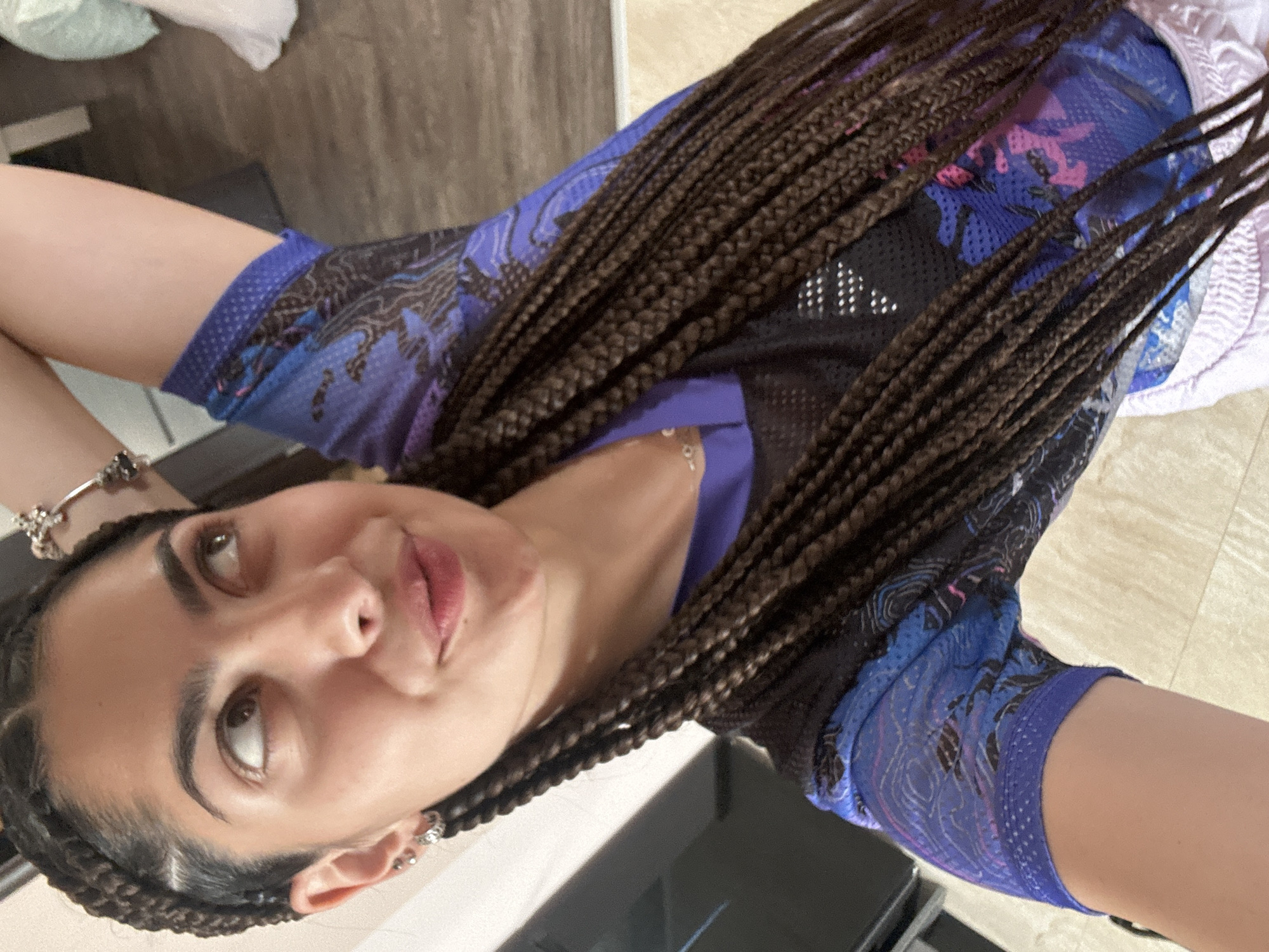 A close-up, high-angle shot shows a young woman with dark brown braids pulled back from her face. She is wearing a blue and black mesh t-shirt with a purple and black design. Her right arm is raised behind her head, and she is looking up and to her left. The background is blurred, but appears to be an indoor setting with light-colored flooring.