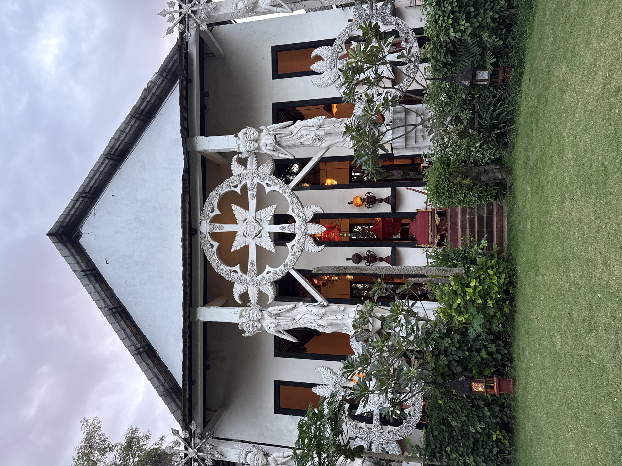 The image depicts the exterior of a building located in an unidentified area of Indonesia. The structure is predominantly white with a gabled roof and features ornate, decorative elements. A large, circular, patterned design dominates the facade, flanked by sculptures of figures. The building has dark-framed windows and an open entrance with glimpses of interior furnishings, including red chairs and lamps. The building's entrance is accessed by a set of steps. Lush greenery surrounds the base of the building, and manicured grass extends to the right of the frame. The sky above is overcast.