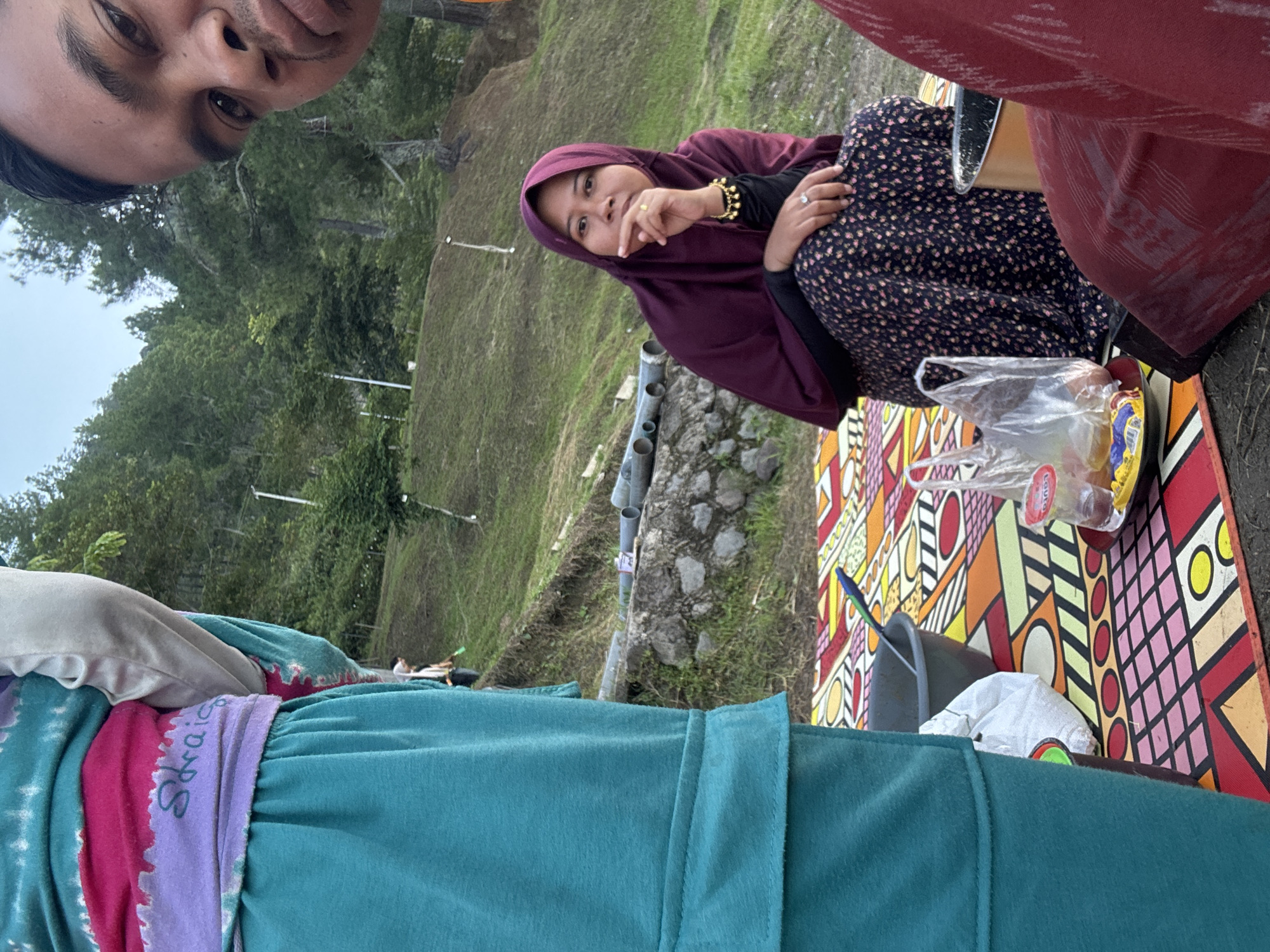 The image depicts a picnic scene outdoors, likely in a hilly or mountainous area, as indicated by the lush green slopes and trees in the background. The location is specified as Kalalengkio, Indonesia.
In the foreground, to the left, a person is partially visible, wearing a teal-colored outfit. A prominent detail on their clothing is the word "Sraic" written in a script across a purple and pink band.
To the right, a man's face is partially visible, taking up the upper right portion of the frame. He appears to be looking towards the camera.
In the middle ground, a woman is seated on a colorful picnic blanket. She is wearing a dark purple hijab and a dark blue floral-patterned skirt. She has a ring on her right ring finger and a bracelet with small dangling charms on her left wrist. She is resting her chin on her hand, looking thoughtfully towards the viewer.
The picnic blanket is laid out on the ground, featuring a vibrant geometric pattern with colors like red, yellow, green, blue, and pink. Several objects are placed on the blanket, including a dark gray bowl with a green spoon, a white plastic bag, a small plate with food items, and a clear plastic container with a red lid. There's also a glimpse of a metallic pot to the right of the woman.
The background shows a grassy slope dotted with trees, suggesting a natural, possibly forested, environment. The sky appears overcast or hazy, indicating it might be late afternoon or early morning. In the distance, there are some grey pipes or conduits visible near the stone embankment.
