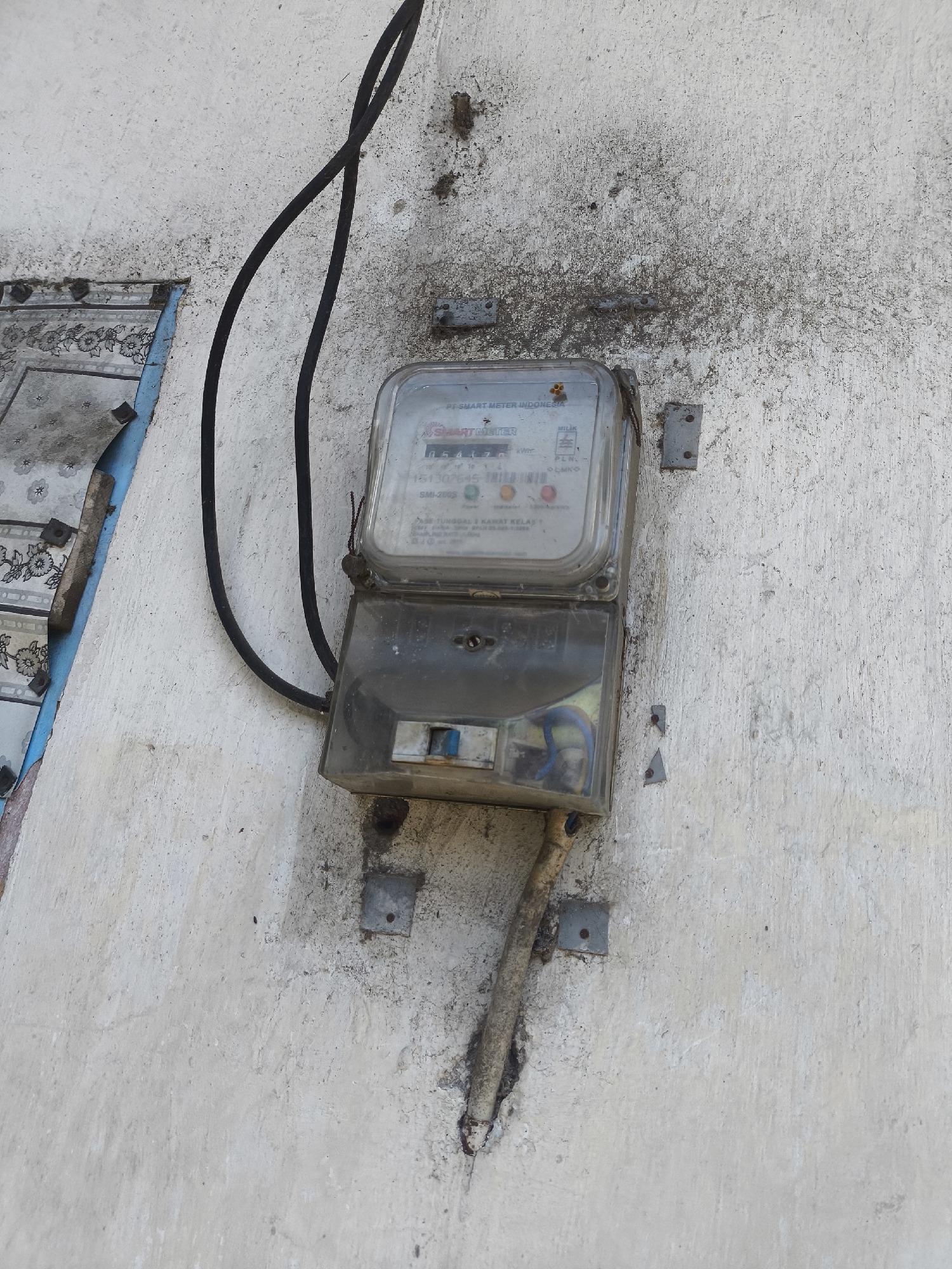 A close-up, high-angle shot captures an outdoor electricity meter mounted on a weathered, off-white concrete wall. The meter, a dual-compartment unit with a clear plastic front, is secured to the wall by four metal brackets. The top compartment displays a digital readout with the text "SMART METER INDONESIA". The bottom compartment shows a blue toggle switch. A black electrical cable, exiting from the left, runs vertically upwards, curving away from the meter. To the left of the meter, a portion of a decorative, patterned wallpaper with floral motifs is visible, peeling away from the wall. The wall itself is marked with grime and water stains, indicating exposure to the elements. The lower portion of the meter unit is connected to a rusted metal conduit that descends diagonally into the wall. The scene suggests an urban or semi-urban setting in Grati, Indonesia, characterized by functional but aging infrastructure.