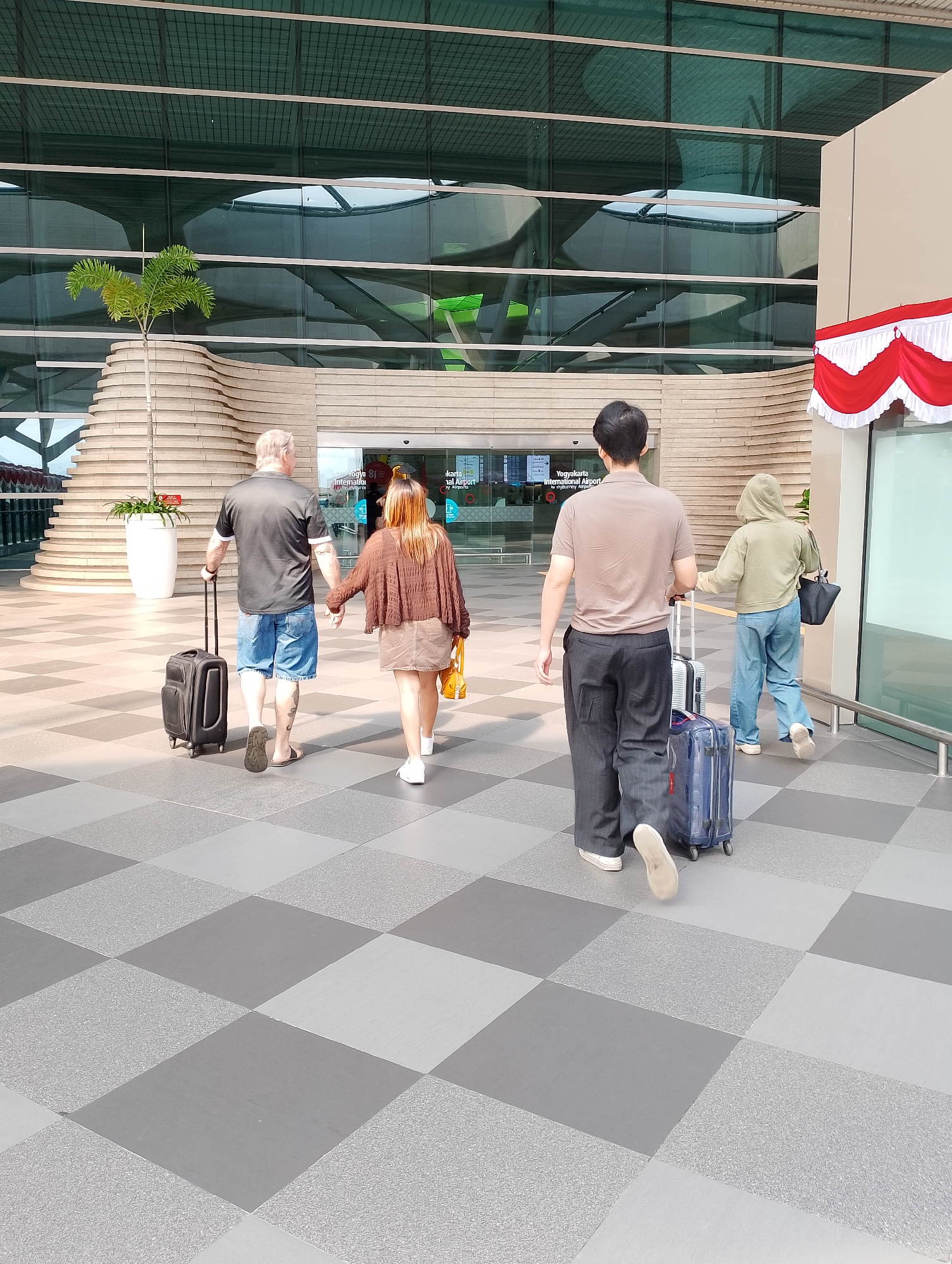 The image depicts four individuals walking away from the camera on the tiled ground outside the Yogyakarta International Airport in Palihan, Indonesia. On the left, an older Caucasian male wearing a black polo shirt and denim shorts is pulling a black suitcase. Beside him, a younger East Asian female with long brown hair, wearing a brown cardigan and a beige skirt, is holding his hand and carrying a yellow bag. To their right, an East Asian male in a tan polo shirt and dark striped pants is pulling a silver suitcase. Behind him, an East Asian female wearing a green hoodie and blue jeans is pulling a blue suitcase and carrying a black tote bag. The airport building features a modern glass facade with visible interior structures and a large, curved architectural element. A small palm tree in a white planter is situated near the entrance. Red and white decorative fabric is visible hanging from the building on the right. The ground is paved with alternating light and dark grey square tiles, with some tiles featuring a textured pattern. The overall scene suggests people arriving at or departing from the airport.