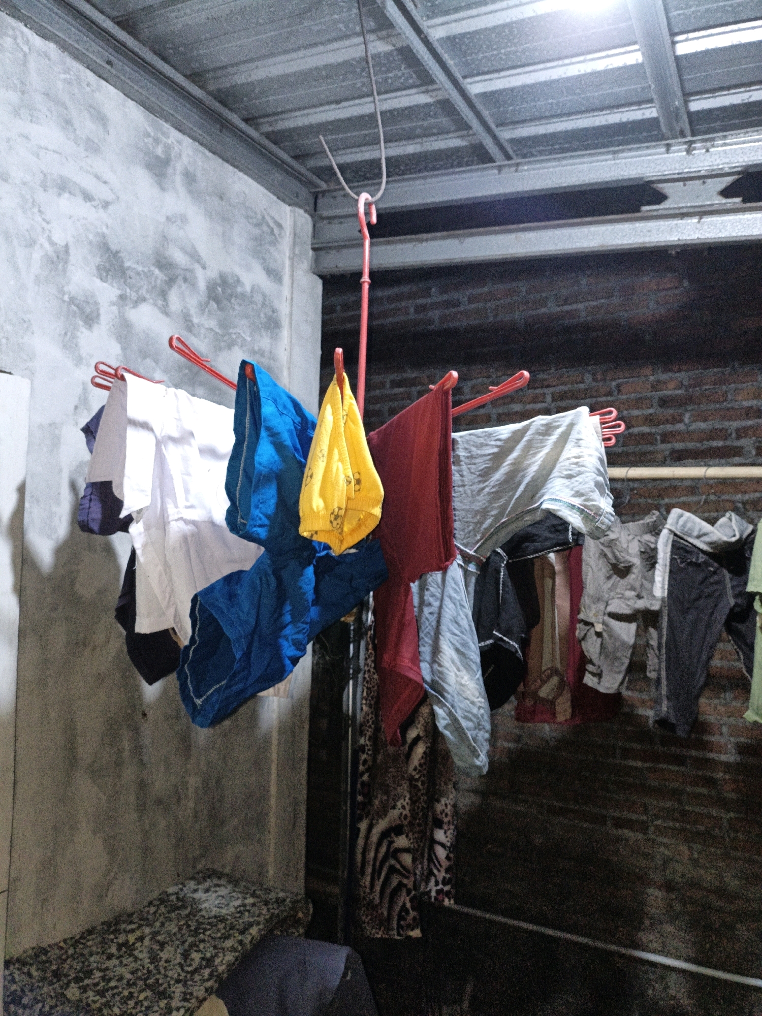 The image depicts an outdoor laundry drying area, likely in Cikal, Indonesia, as indicated by the context. A red clothes drying rack is suspended from a metal ceiling structure, holding a variety of garments. These include white shirts, blue shorts, a yellow garment with a football print, and a red t-shirt. Other clothing items are visible on a second, lower clothesline, such as grey pants, a leopard print garment, and other assorted fabrics. The background consists of a textured concrete wall on the left and a brick wall on the right, with a corrugated metal roof overhead. A bright light source is visible in the upper center of the image. The overall scene suggests a domestic setting where laundry is being air-dried.