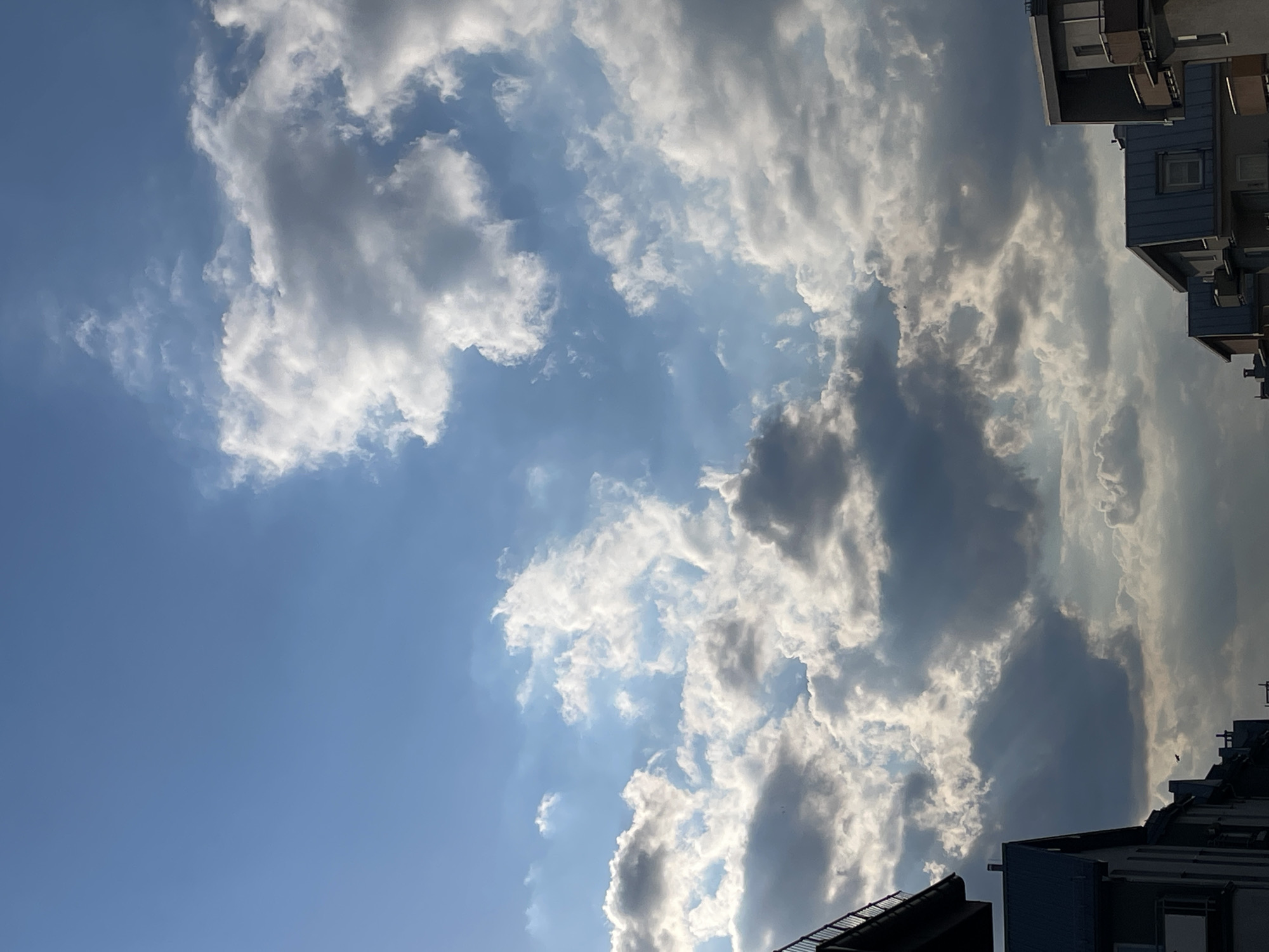 The image shows a view of the sky, partially obscured by buildings in an urban area. The sky is blue with cumulus clouds. The buildings are multi-story and appear to be residential. The location is in the city of Unknown, Unknown.