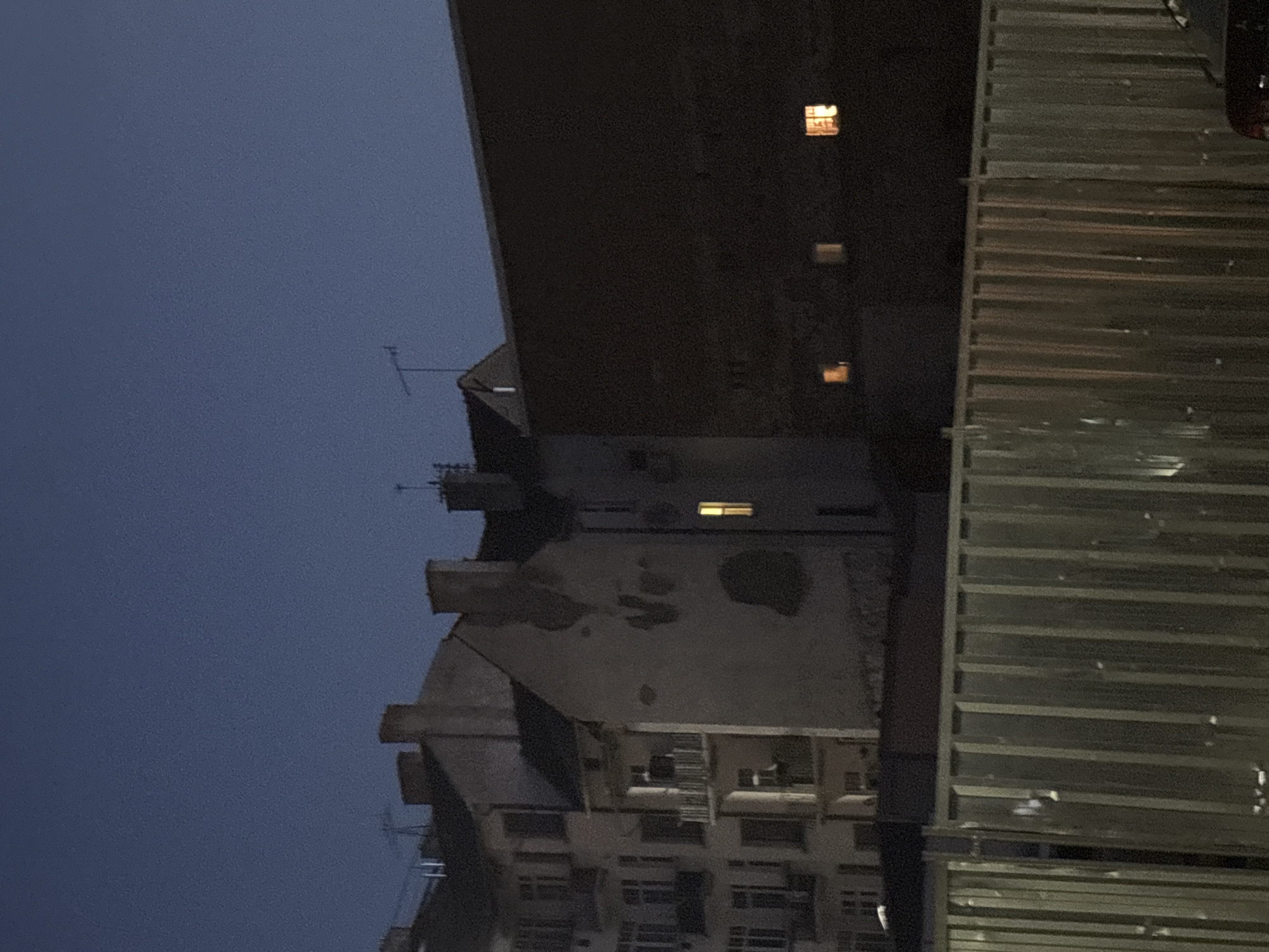 The image depicts an urban scene at dusk or night. In the foreground, a corrugated metal fence runs horizontally across the frame. Behind the fence, several buildings are visible. The leftmost building appears to be a multi-story apartment complex with balconies and windows. Adjacent to it is a taller, older building with textured walls, showing signs of wear and possibly damage. Further to the right, a dark, imposing structure with a flat roof dominates a significant portion of the frame. Several windows are illuminated in the buildings, suggesting occupancy. The sky is a deep blue, transitioning to darkness. The overall atmosphere is one of quietude and urban isolation.