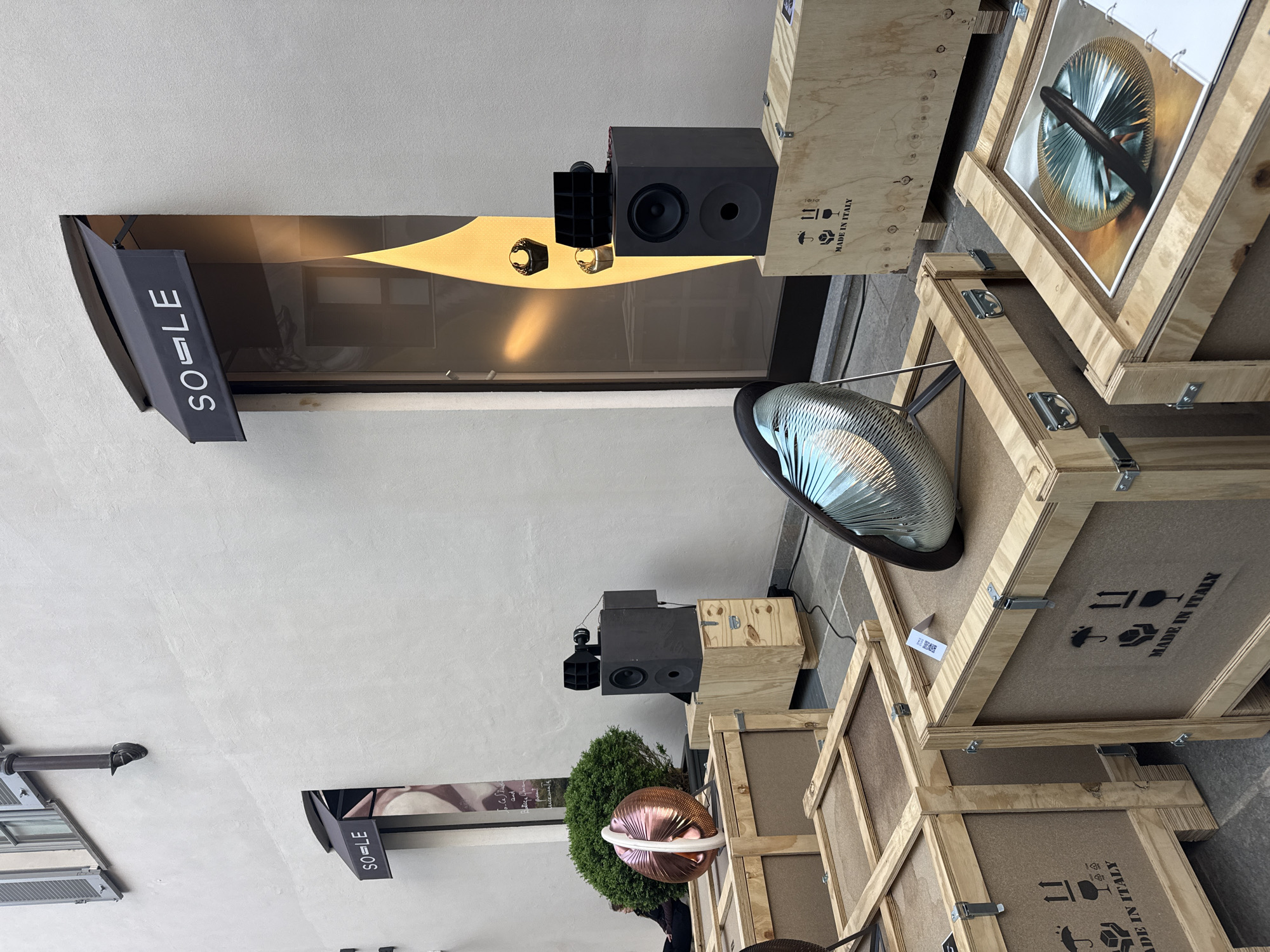 The image captures an outdoor display or art installation on an urban street, likely in Milan, Italy, under bright daylight. The primary subjects are an arrangement of wooden shipping crates, professional audio equipment, and decorative elements.
Numerous light-colored wooden crates are stacked and placed on a paved surface in the foreground and midground. Many of these crates are stenciled with "MADE IN ITALY" in black text, accompanied by international shipping symbols such as a fragile glass, an umbrella (keep dry), and "this way up" arrows. One crate displays an image of a circular, spoked metallic object.
Interspersed with the crates are two prominent black audio speakers, one featuring an additional gridded horn component. These speakers are positioned on smaller wooden bases or crates. Decorative items include a shiny, corrugated silver dome-like sculpture, a metallic sphere (appearing copper or bronze) adorned with a white band, and a small, vibrant green bush.
In the background, a light-colored building facade is visible, featuring a large storefront window that reflects its surroundings. Above the window, two black awnings or banners are extended, each displaying the white sans-serif text "SO-LE". Another "SO-LE" sign is visible further down the street on the building. The overall setup suggests a temporary exhibition, retail display, or a public art installation. No people are clearly visible in the frame. The bright lighting indicates a sunny day with clear weather.