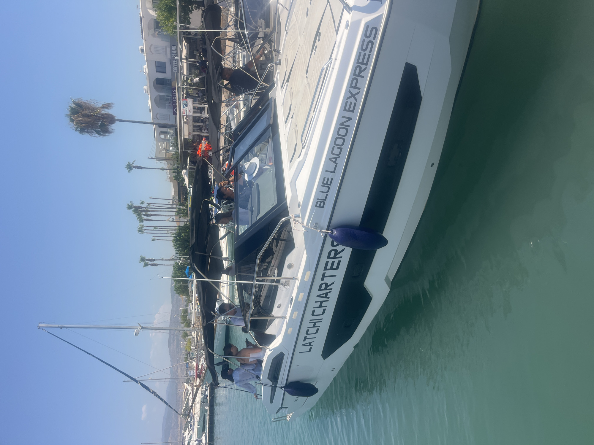 The image shows a white motorboat named "LATCHI CHARTERS BLUE LAGOON EXPRESS" docked in a marina. The boat is white with black accents and has several people on board, some standing and some seated. The people appear to be enjoying a sunny day. The background shows a row of buildings with palm trees in front, suggesting a coastal location. The water is a clear turquoise color. Based on the context of the boat's name and the surrounding environment, this scene is likely in the Latchi area of Polis Chrysochous, Cyprus, a popular destination for boat tours and water activities.
