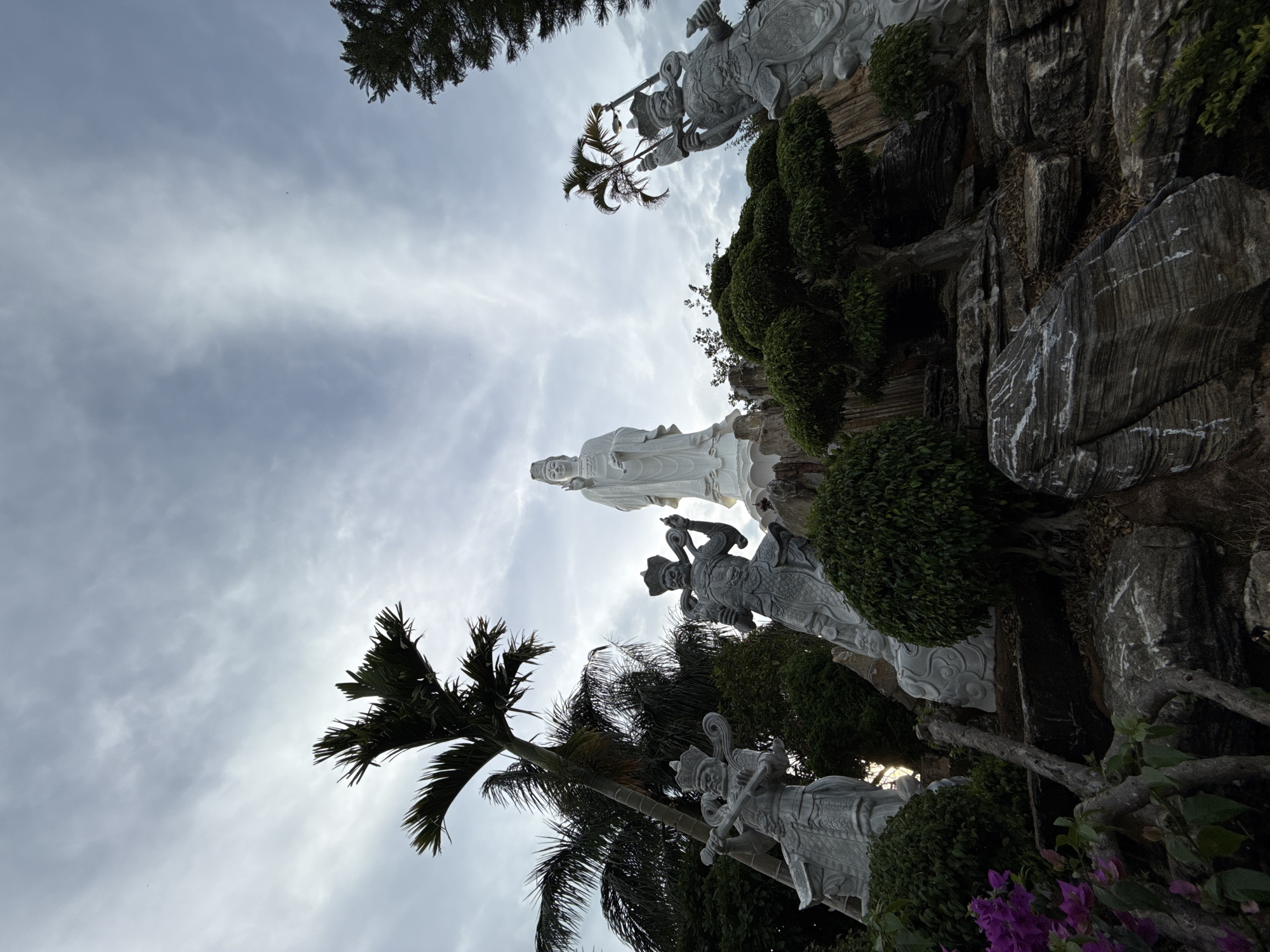 The image shows a low-angle view of a complex of statues and landscaping in Thôn 5, Vietnam. A large white statue of a female figure stands prominently, flanked by smaller, intricately carved stone statues of warrior figures in traditional attire. These figures appear to be part of a rock garden featuring greenery and rocky formations. Palm trees and other vegetation frame the scene. The sky is overcast with diffused sunlight filtering through.